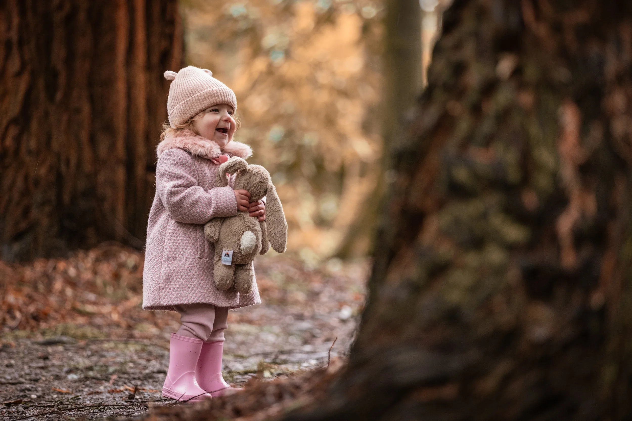 A smiling young girl in pink rain boots and a pink coat, holding a plush bunny, stands in a wooded area during fall.