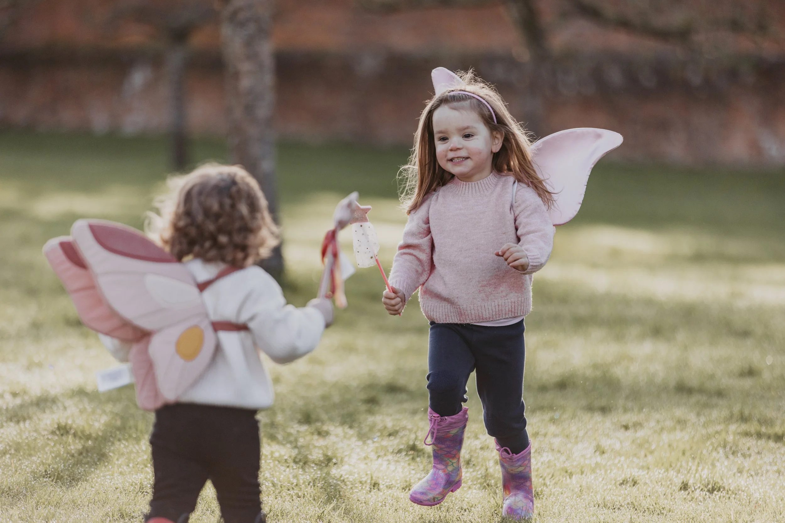 Two young girls with fairy wings playing outdoors on grass, one girl is running holding a wand with a star on top, the other girl is standing with butterfly wings, wearing a pink sweater and rain boots.