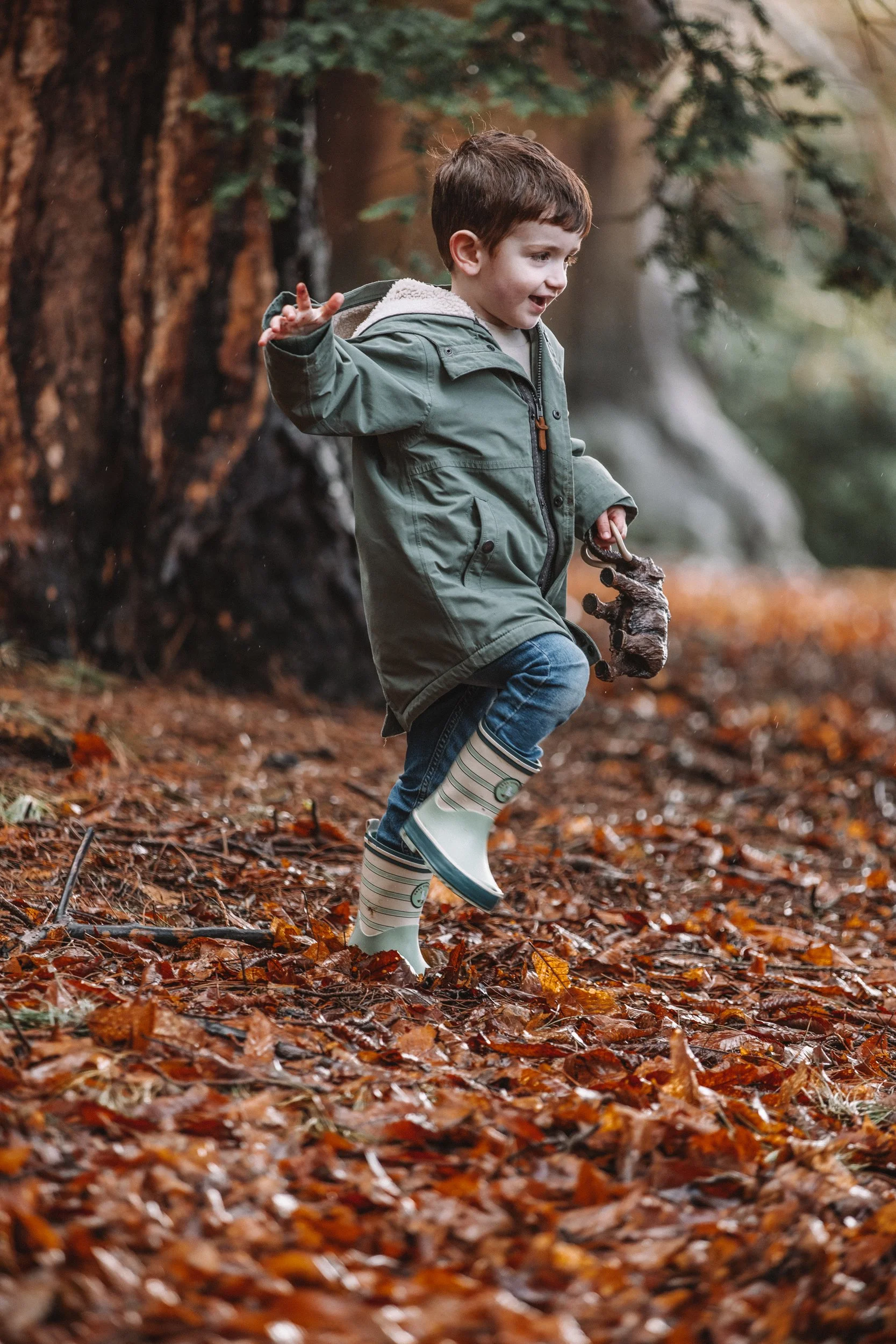 A young boy in an olive green raincoat and striped rubber boots playing in a forest covered with fallen autumn leaves, holding a small animal or toy.