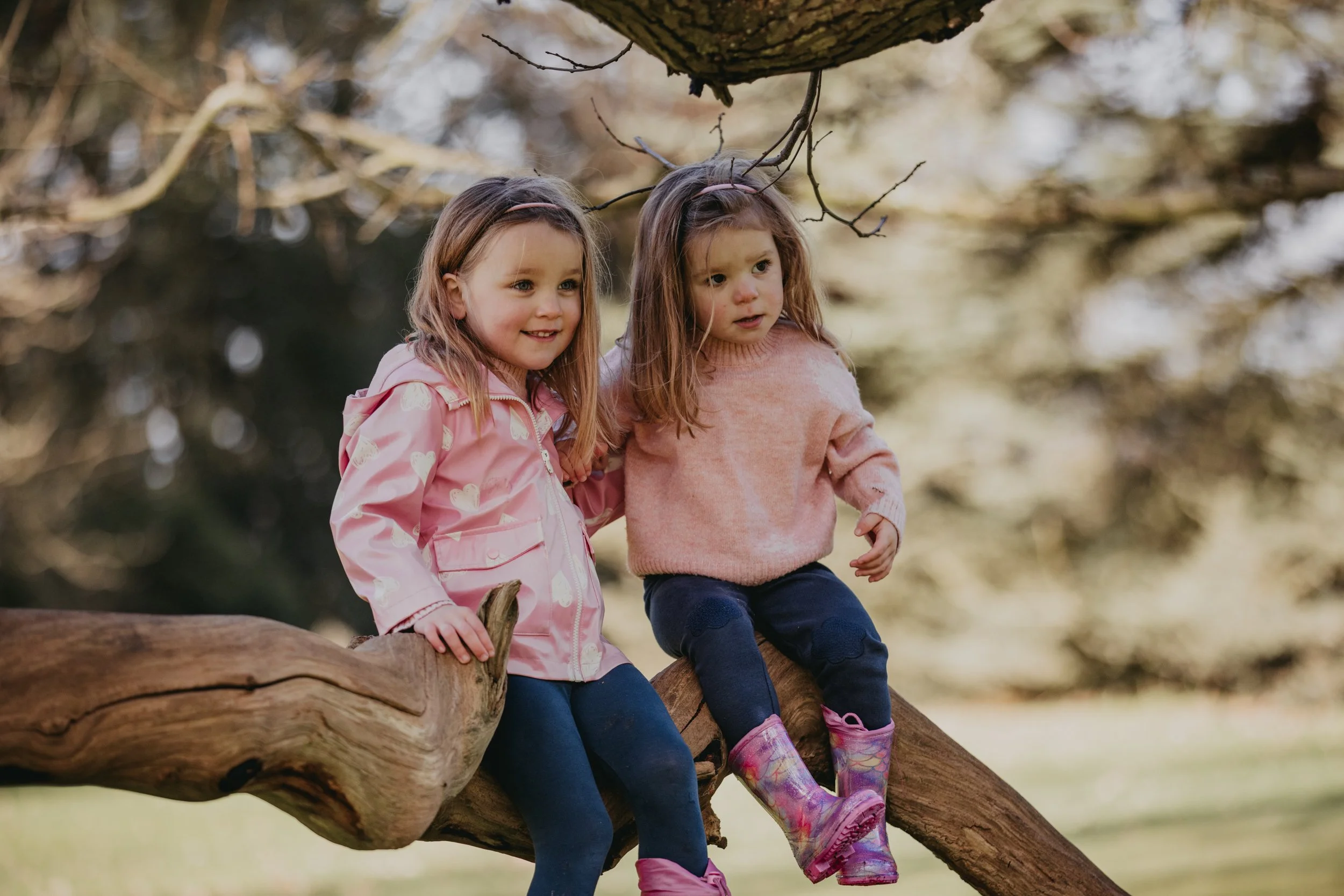Two young girls sitting on a tree branch outdoors with a blurred forest background.