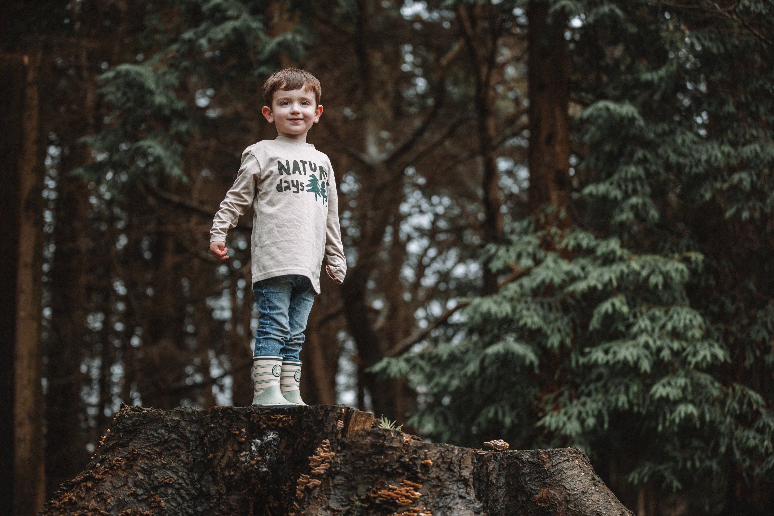 A young boy standing on a large tree stump in a forest, wearing a beige long-sleeve shirt and rain boots.