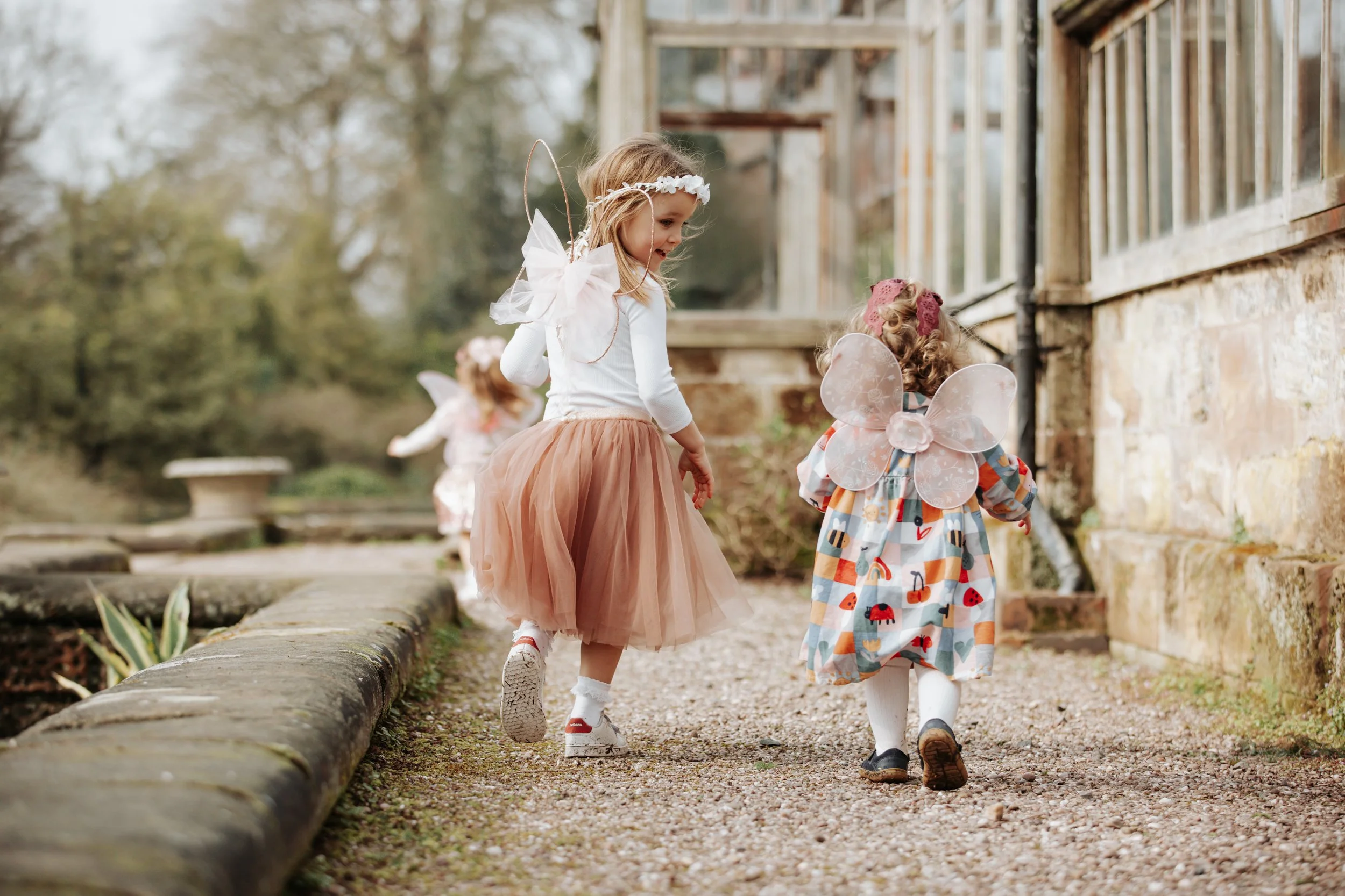 Three young girls dressed as fairies in colorful costumes walking outdoors along a stone pathway near a rustic building with a stone wall and large windows, surrounded by trees.