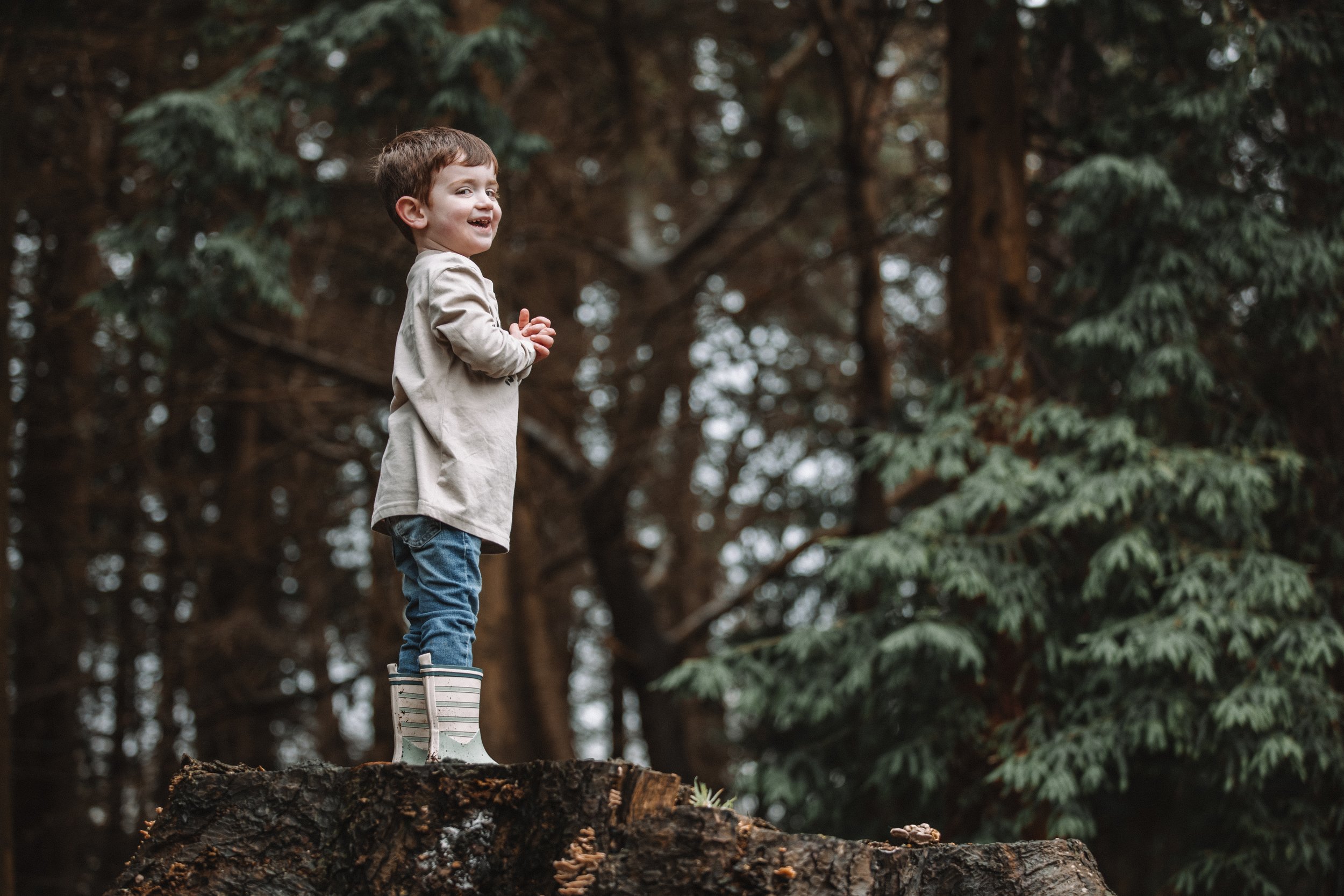 a young boy standing on a tree stump in a forest, wearing rain boots, a beige jacket, and jeans, smiling and looking to the side.