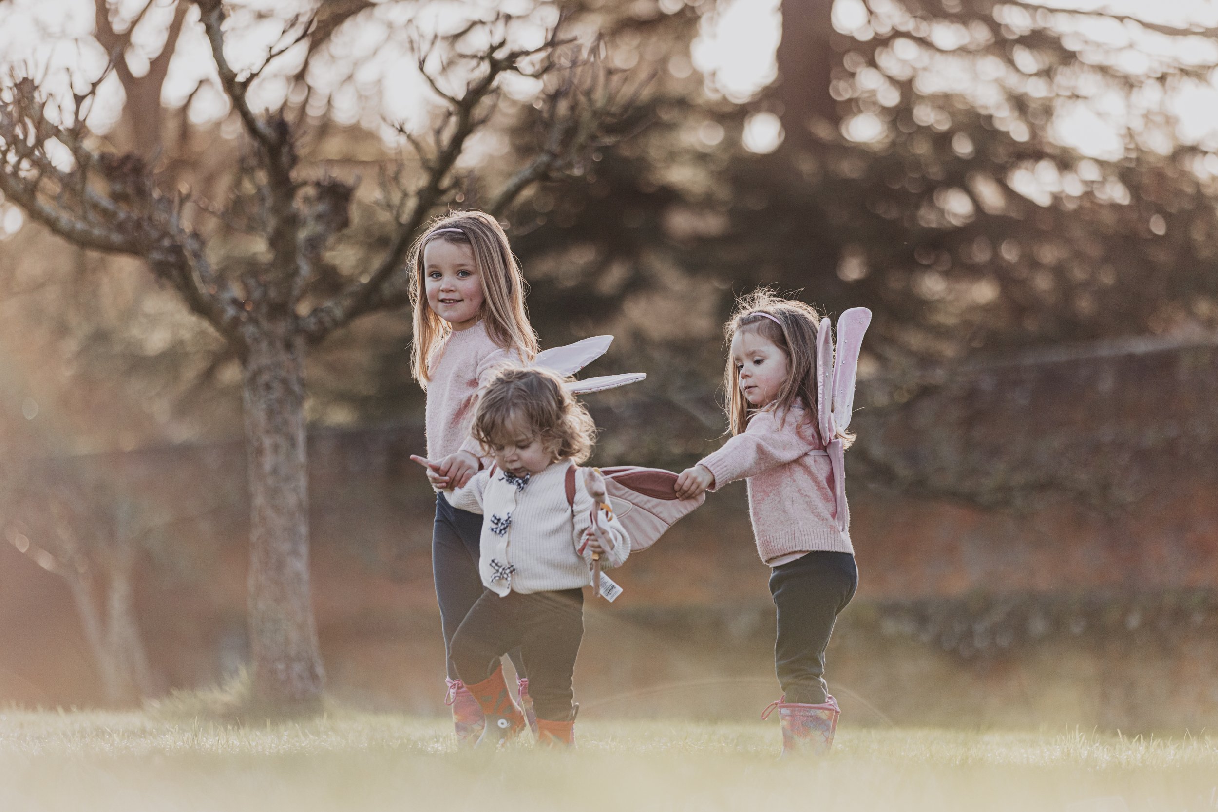 Three young girls with fairy wings playing outdoors on a grassy field with trees in the background during sunset.