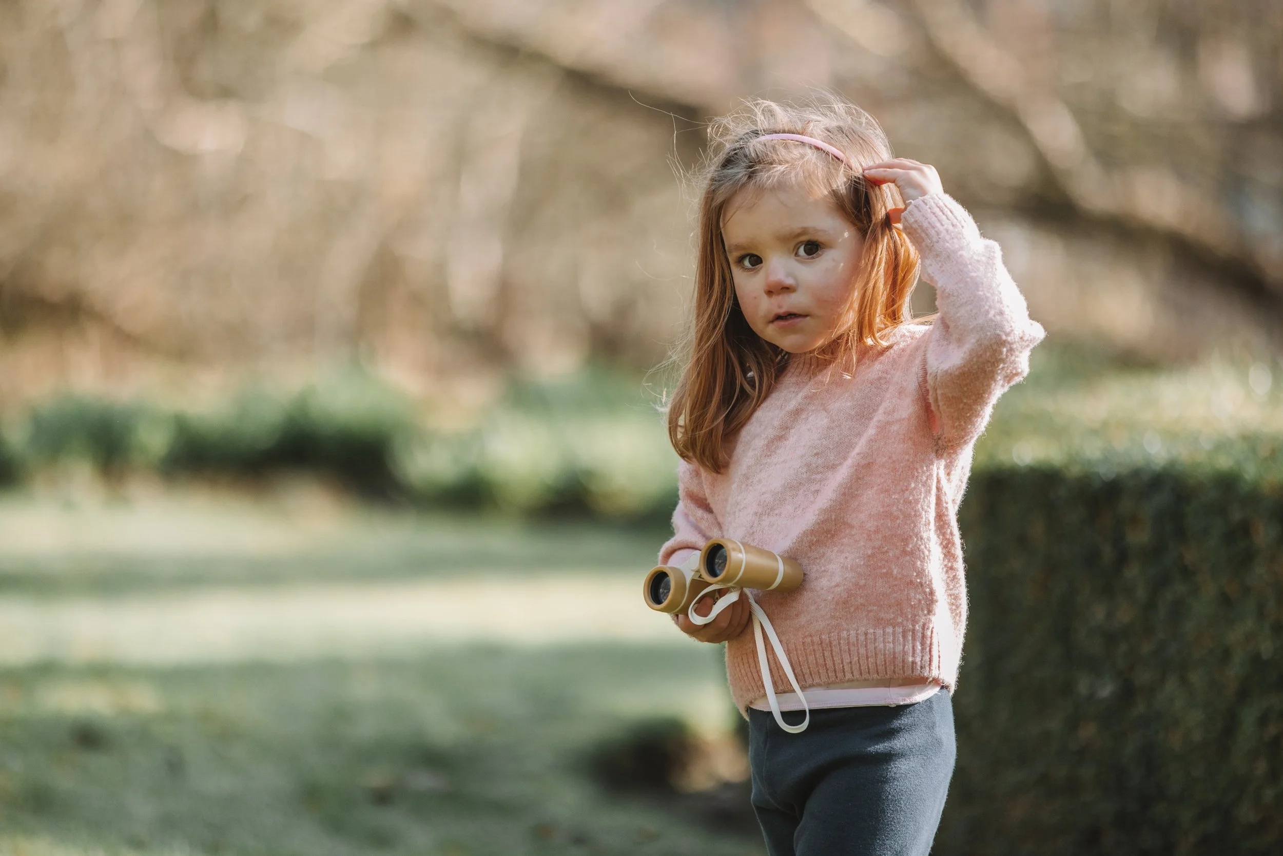 A young girl with red hair and light skin, wearing a pink sweater and dark pants, is outdoors in a park. She is holding binoculars in one hand and touching her head with the other. The background shows blurred trees and greenery.