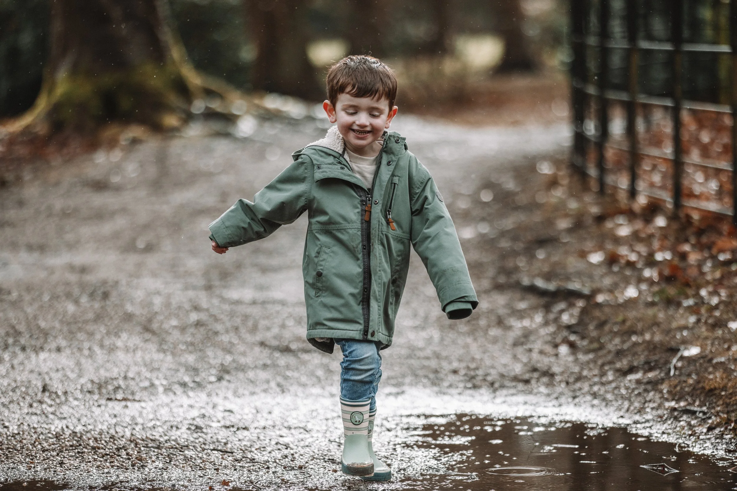 A young boy in a green raincoat and rain boots walking through a muddy puddle on a trail in a forest during daytime.