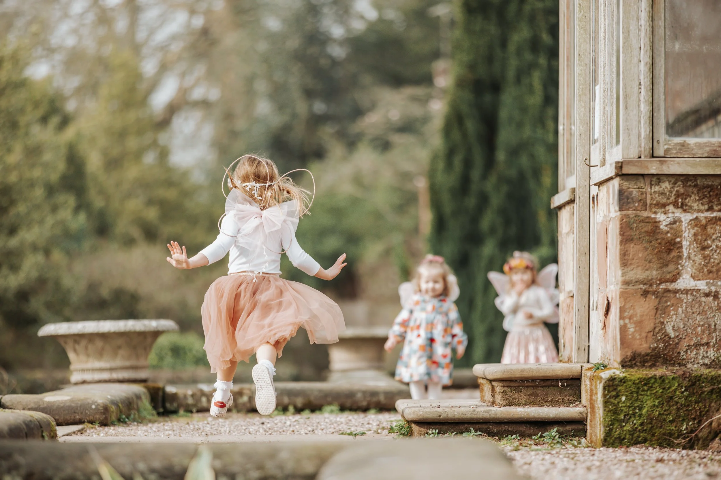 Three young girls dressed as fairies playing outdoors, one girl with a tan tutu running towards two girls dressed in floral dresses with fairy wings near a brick building.