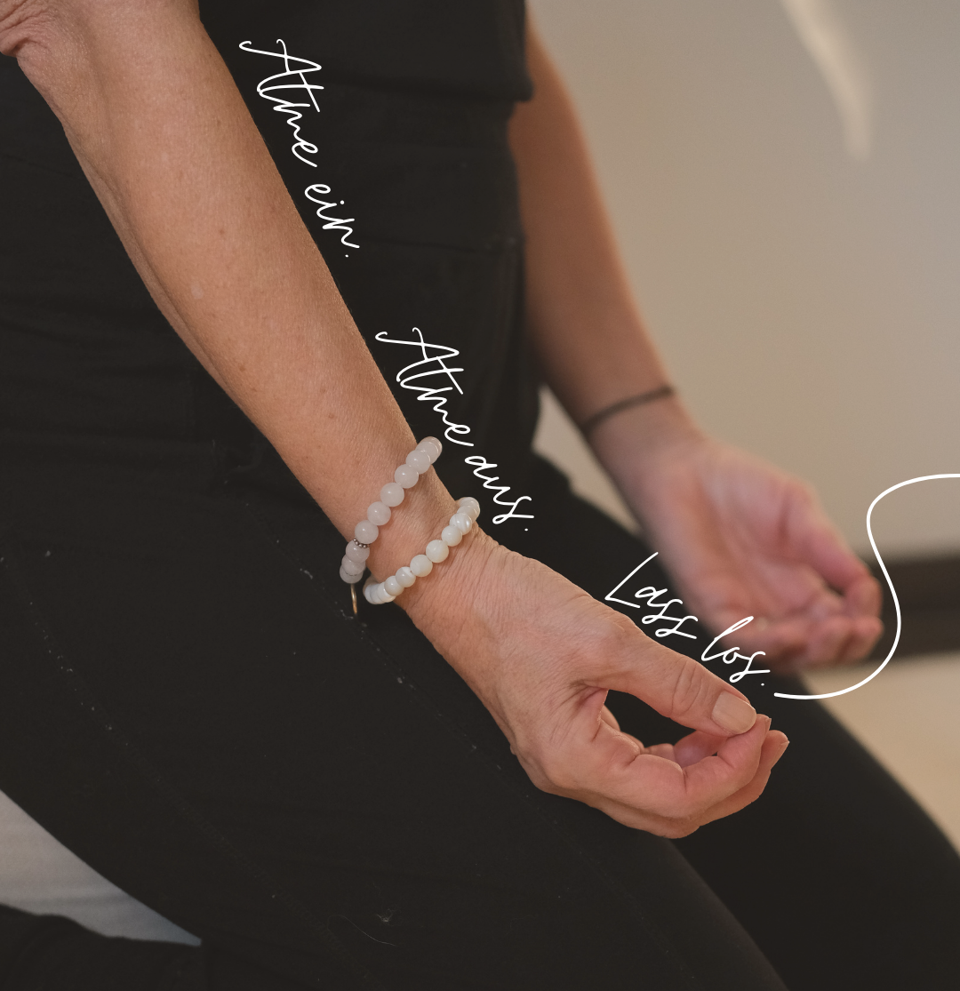 Nahaufnahme einer Person, die im Lotossitz meditieren macht, mit weißen Perlenarmbändern am Handgelenk, das an den Knien ruht.