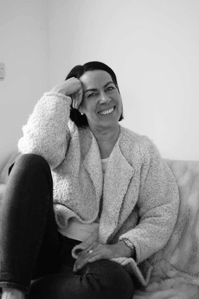 Eine lachende Frau mit dunklem Haar, die auf einem Sofa sitzt, den Kopf leicht zur Seite geneigt und die Hand an der Stirn hat, in einem gemütlichen Zimmer.