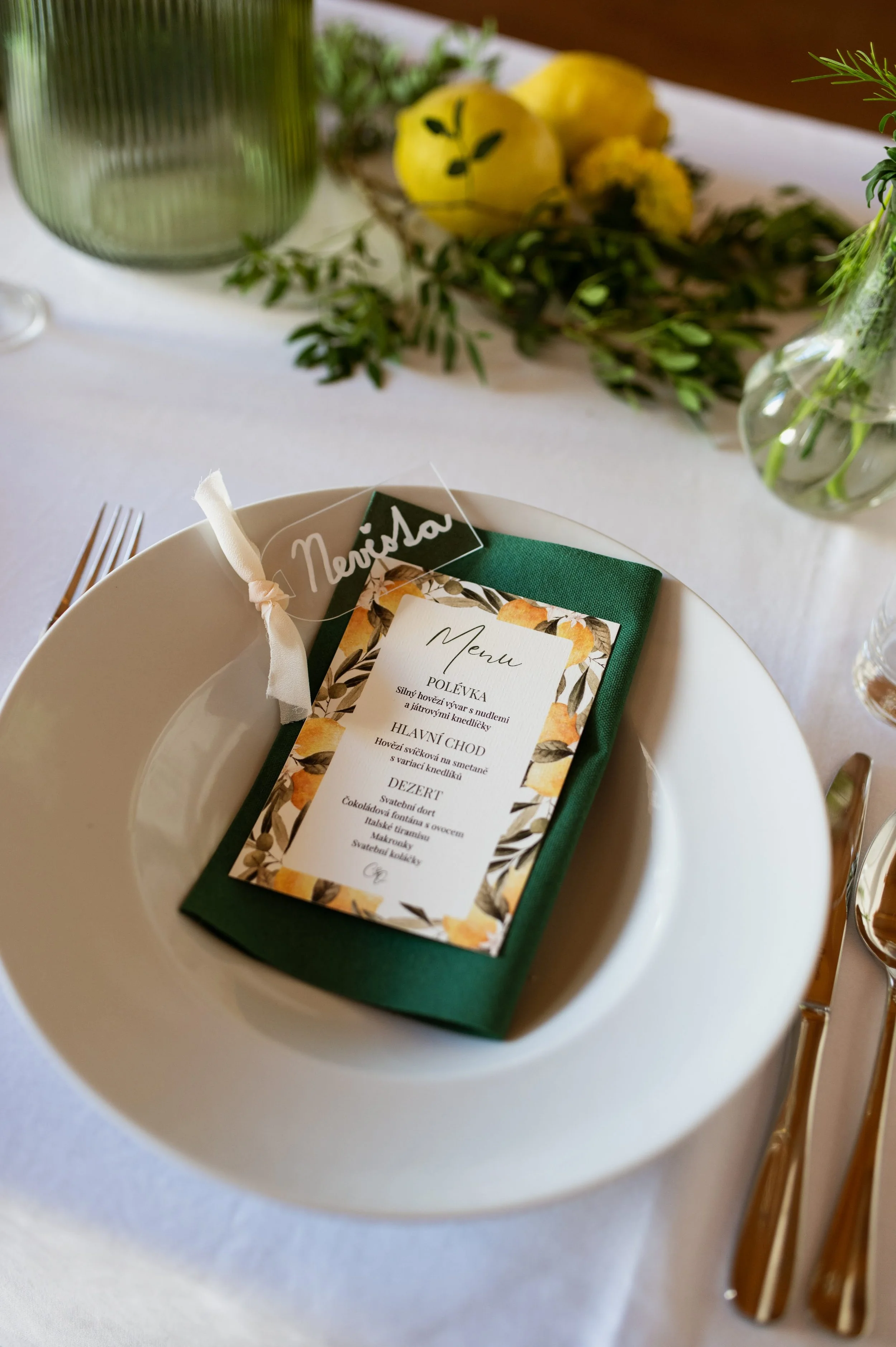 Place setting with a white plate, green napkin, and a menu card with a botanical border, on a table with cutlery, glassware, lemons, and greenery.