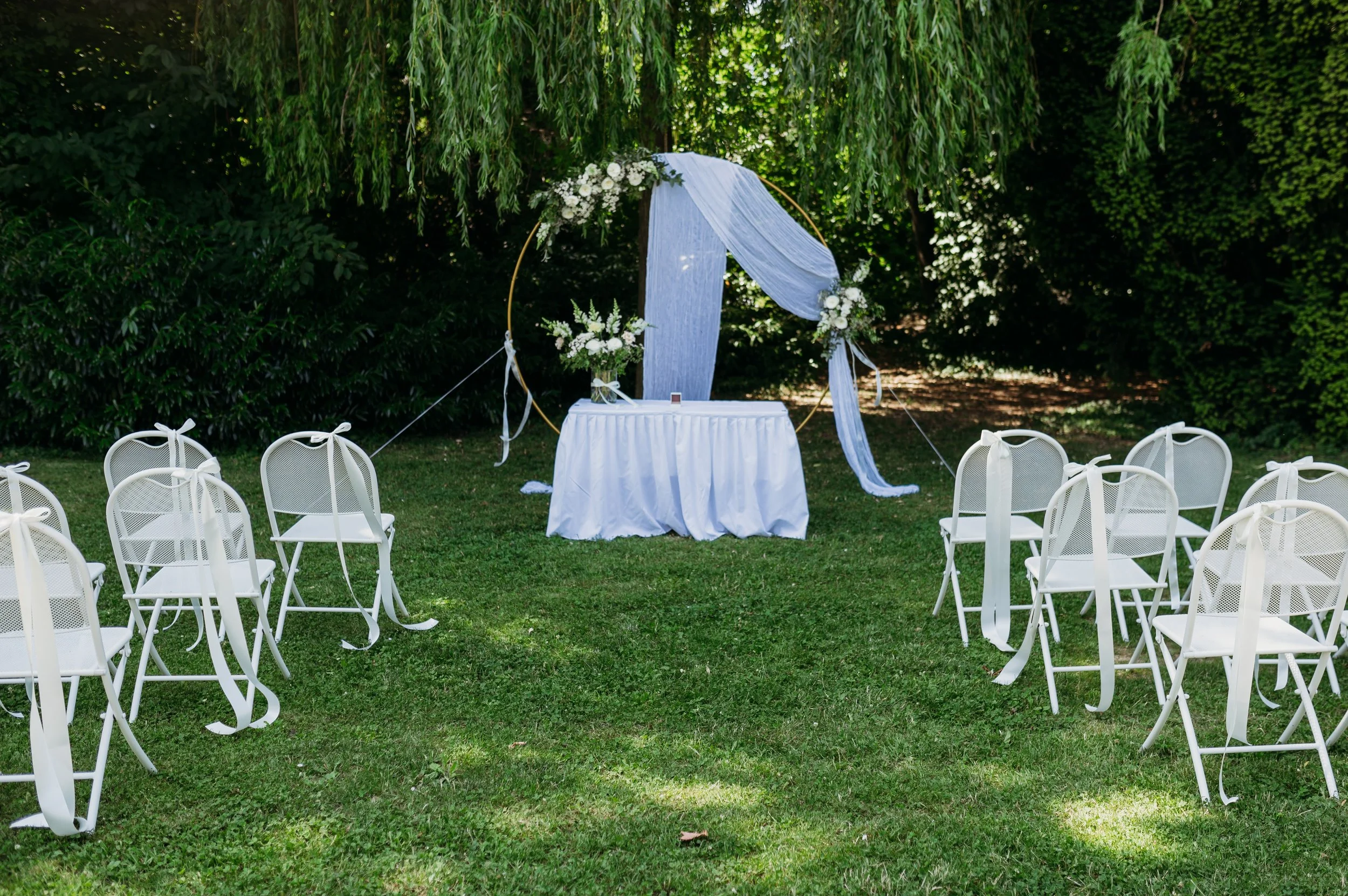 Outdoor wedding setup with white chairs tied with ribbons, a table with a white cloth, and floral arrangements under a decorative hoop, surrounded by greenery.