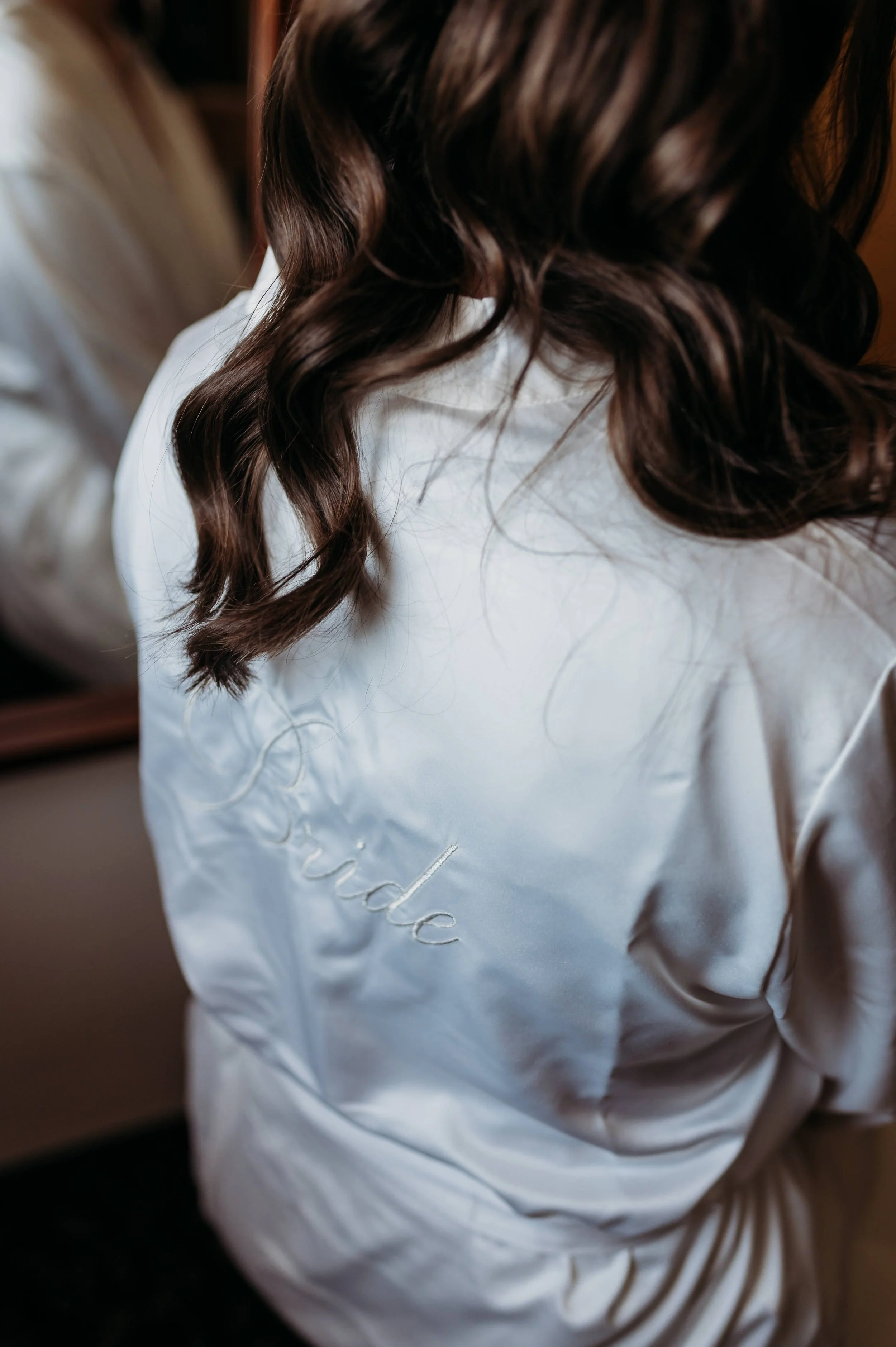 Nevěsta se chystá na obřad v županu s nápisem bride. Hnědé vlnité vlasy.