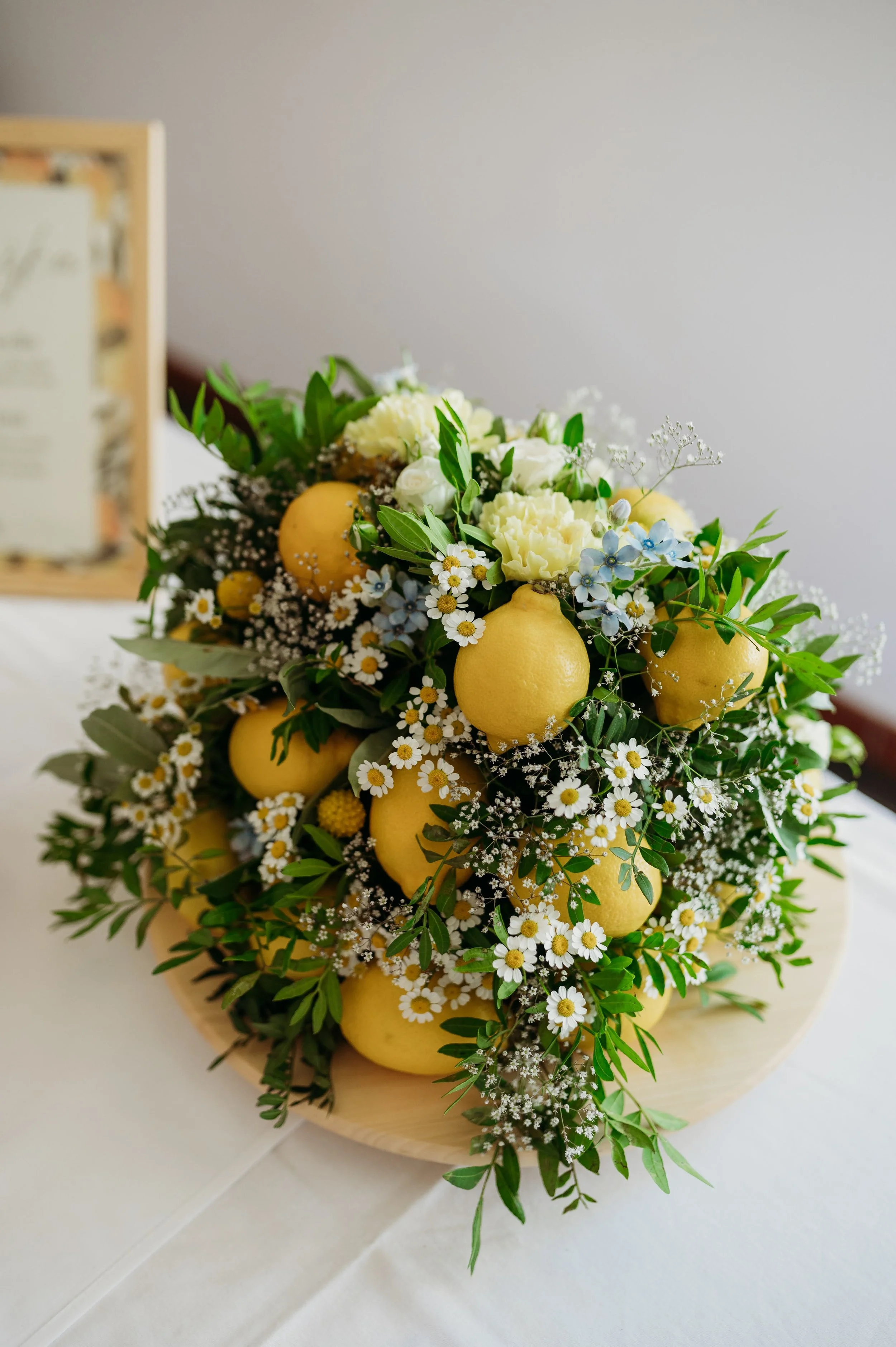 A bouquet of yellow lemons, white flowers, and green foliage arranged in a round wooden tray.