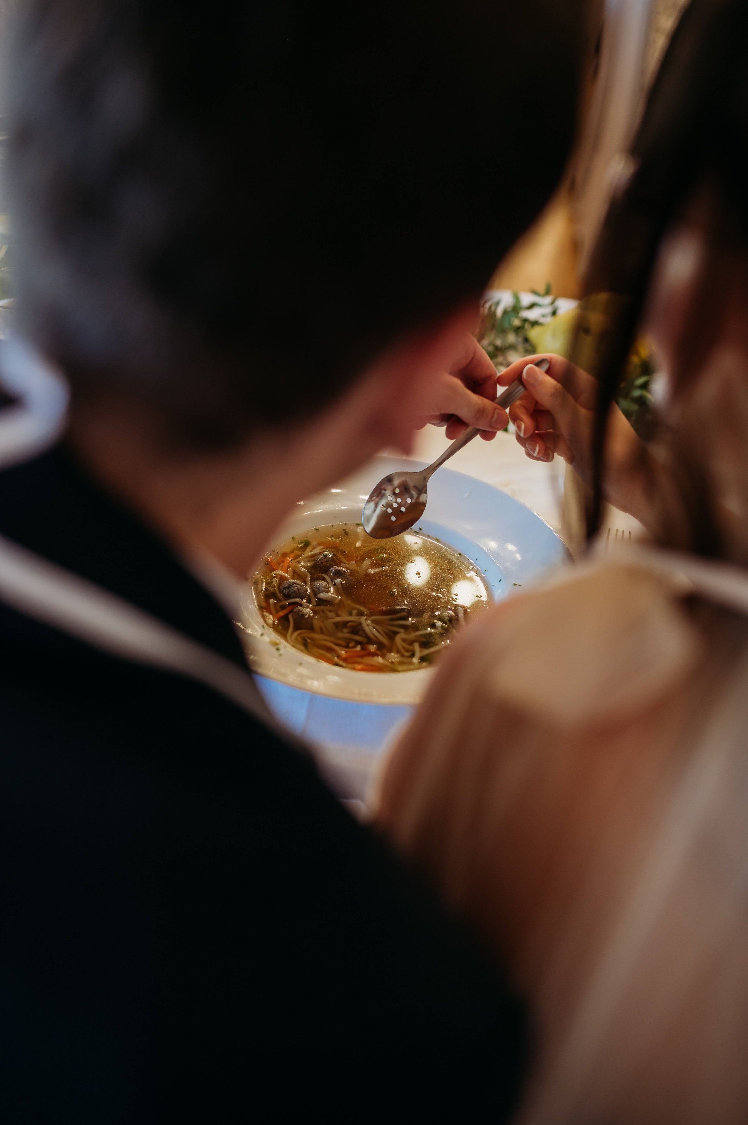A couple eating noodle soup at a table with a bowl of broth and noodles visible. The photo is taken from behind the couple, focusing on their heads and hands.