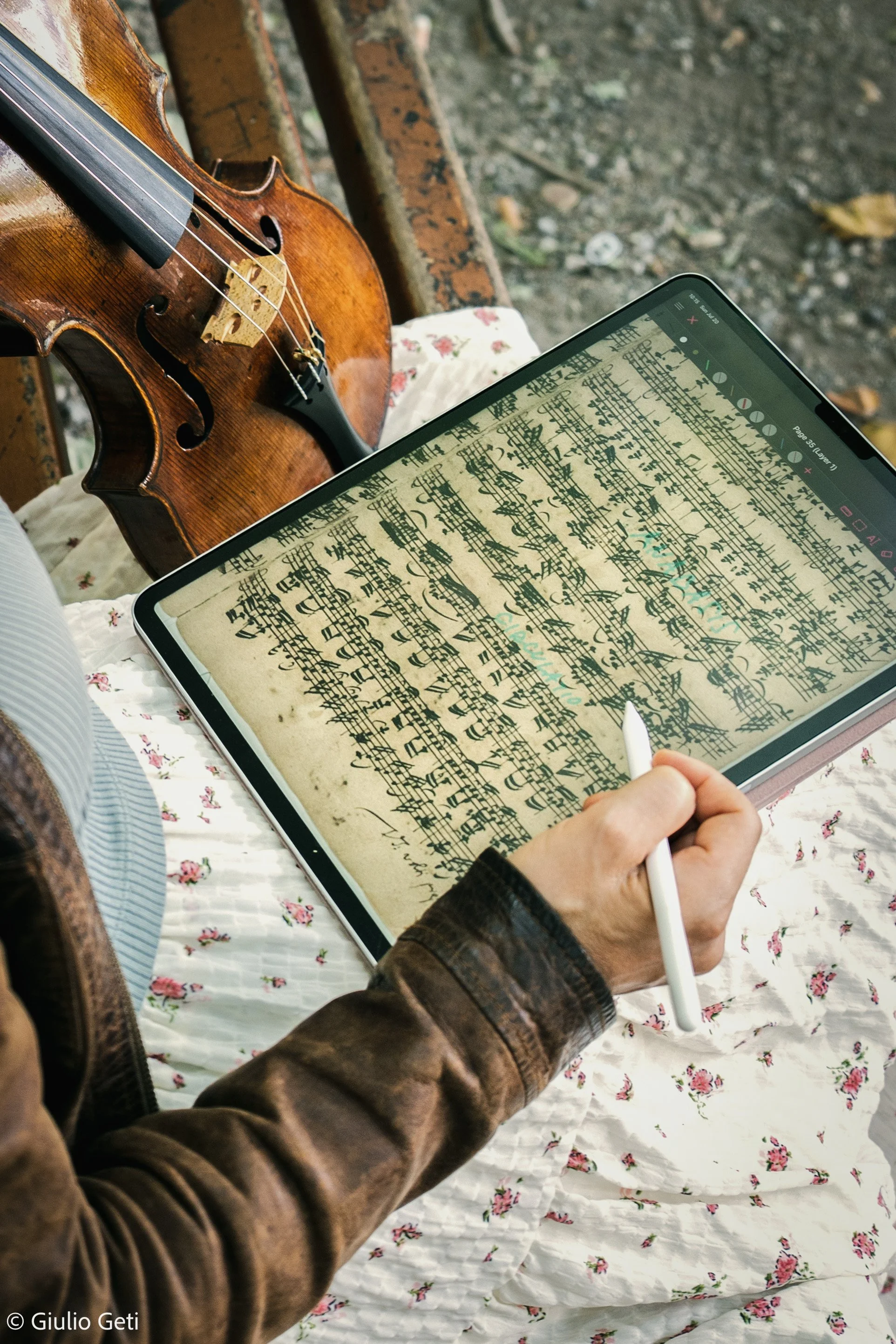 Laura Corolla holding a stylus and editing sheet music on a tablet, with a violin resting nearby on a wooden bench.