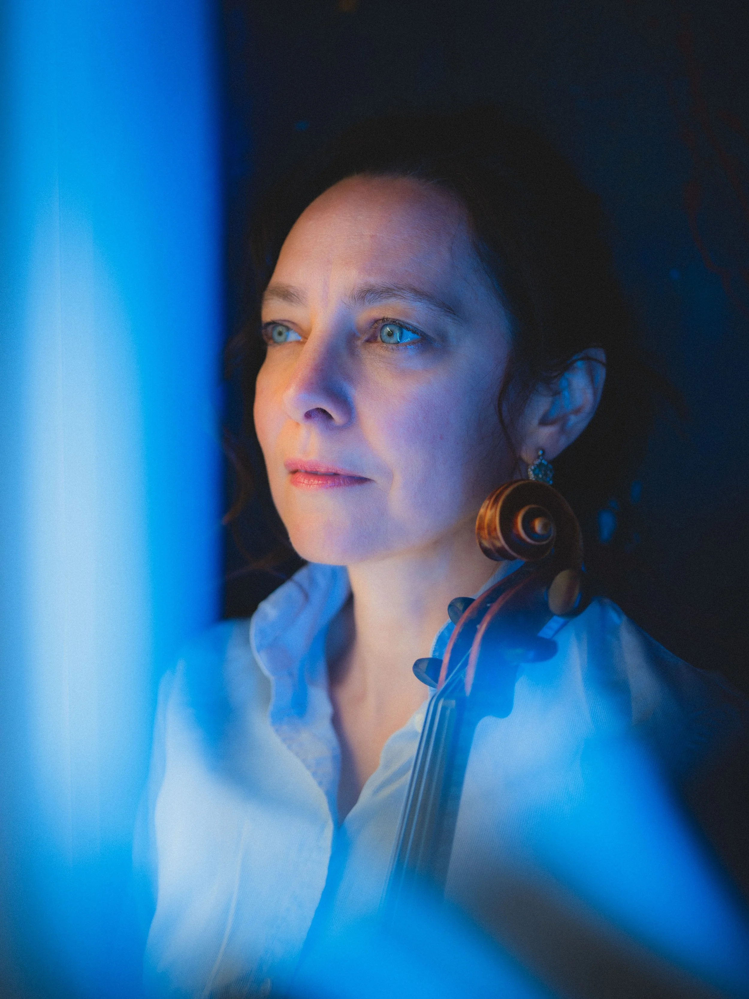 Violinist Laura Corolla,  with blue eyes and dark hair, holds a violin, with blue lighting illuminating her face and a dark background.