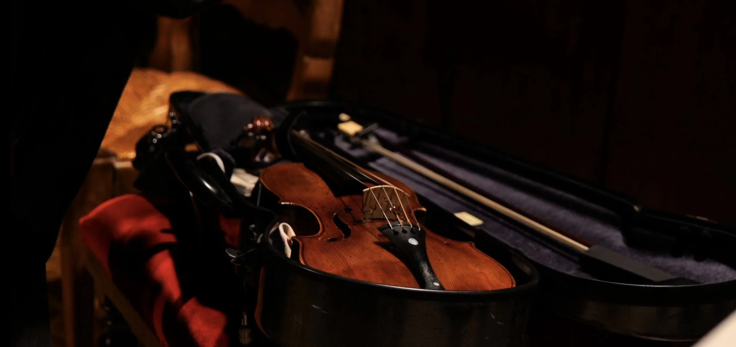 A violin inside an open black case, with the case lid open and some bows inside, resting on a wooden surface.