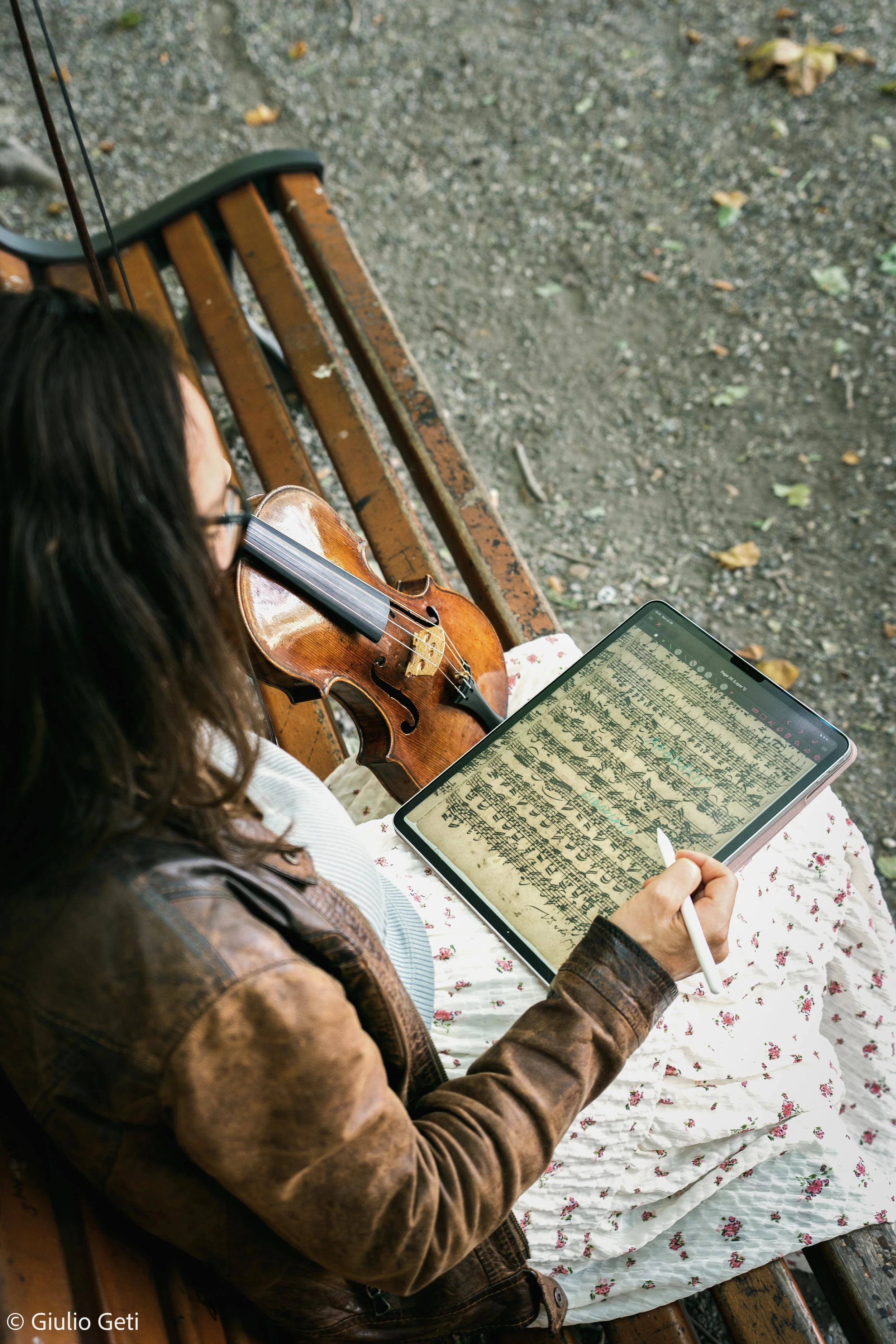 Violinist Laura Corolla, in a brown leather jacket ,sitting on a wooden park bench, holding a tablet with digital musical notation. The ground is gravel with some fallen leaves.