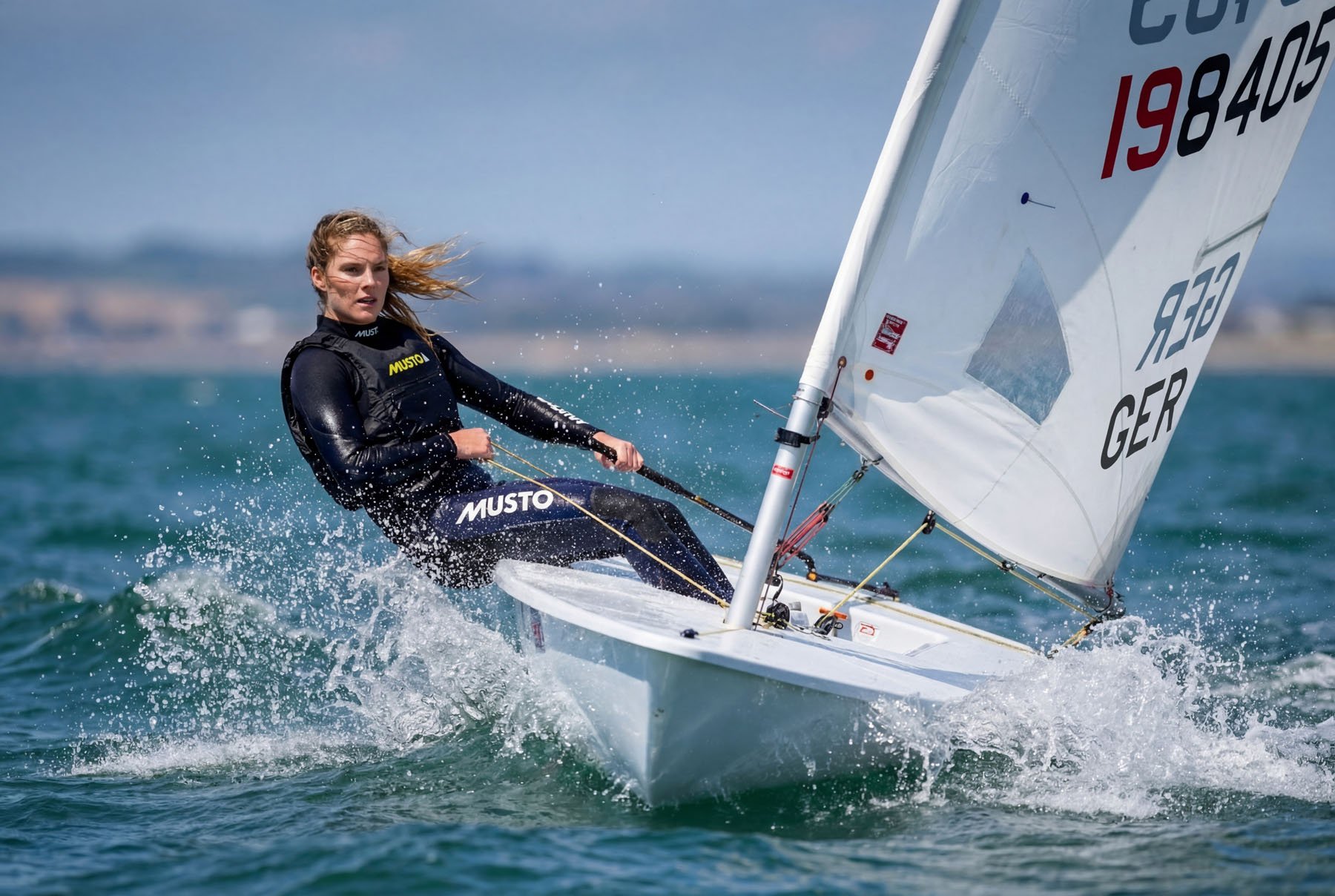 A female sailor on a small sailboat racing on the water, wearing a black Musto wetsuit, with wind blowing her hair as she navigates at high speed.