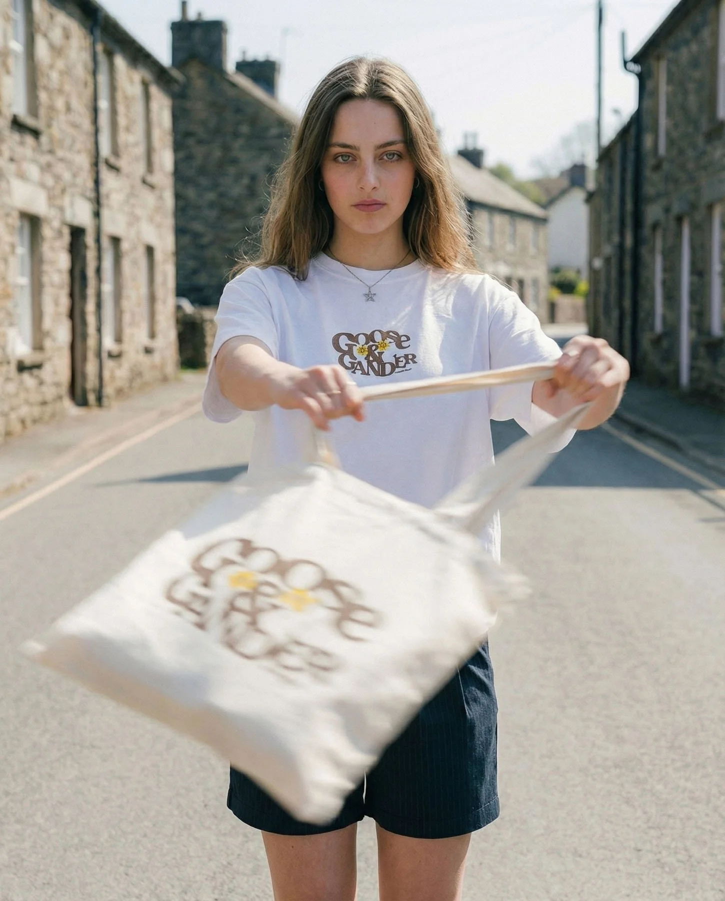 A young woman with long brown hair standing on a street during daytime, holding a tote bag with a design and text that says 'Goose Gander' and a daisy flower in the center, wearing a white t-shirt with a similar logo.