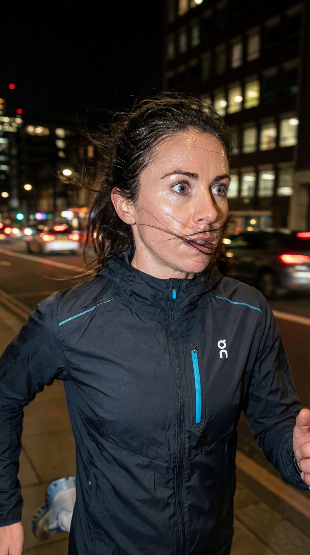 A woman running at night on a city street, wearing a black athletic jacket, with city lights and traffic in the background, her hair blowing in the wind.