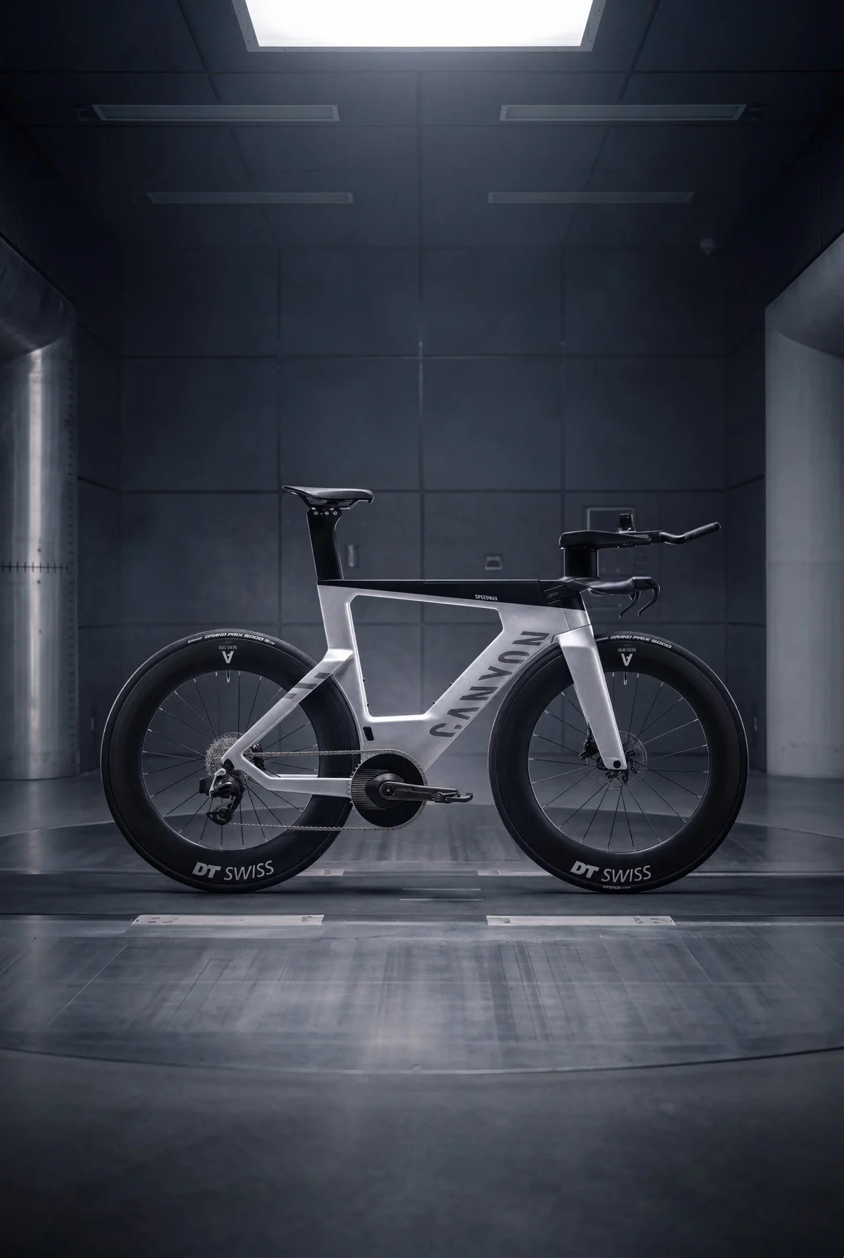 A sleek, modern Canyon Speedmax CFR 1by AXS triathlon bicycle in a dark, industrial-looking wind tunnel room with metal and concrete walls, illuminated by a ceiling light.