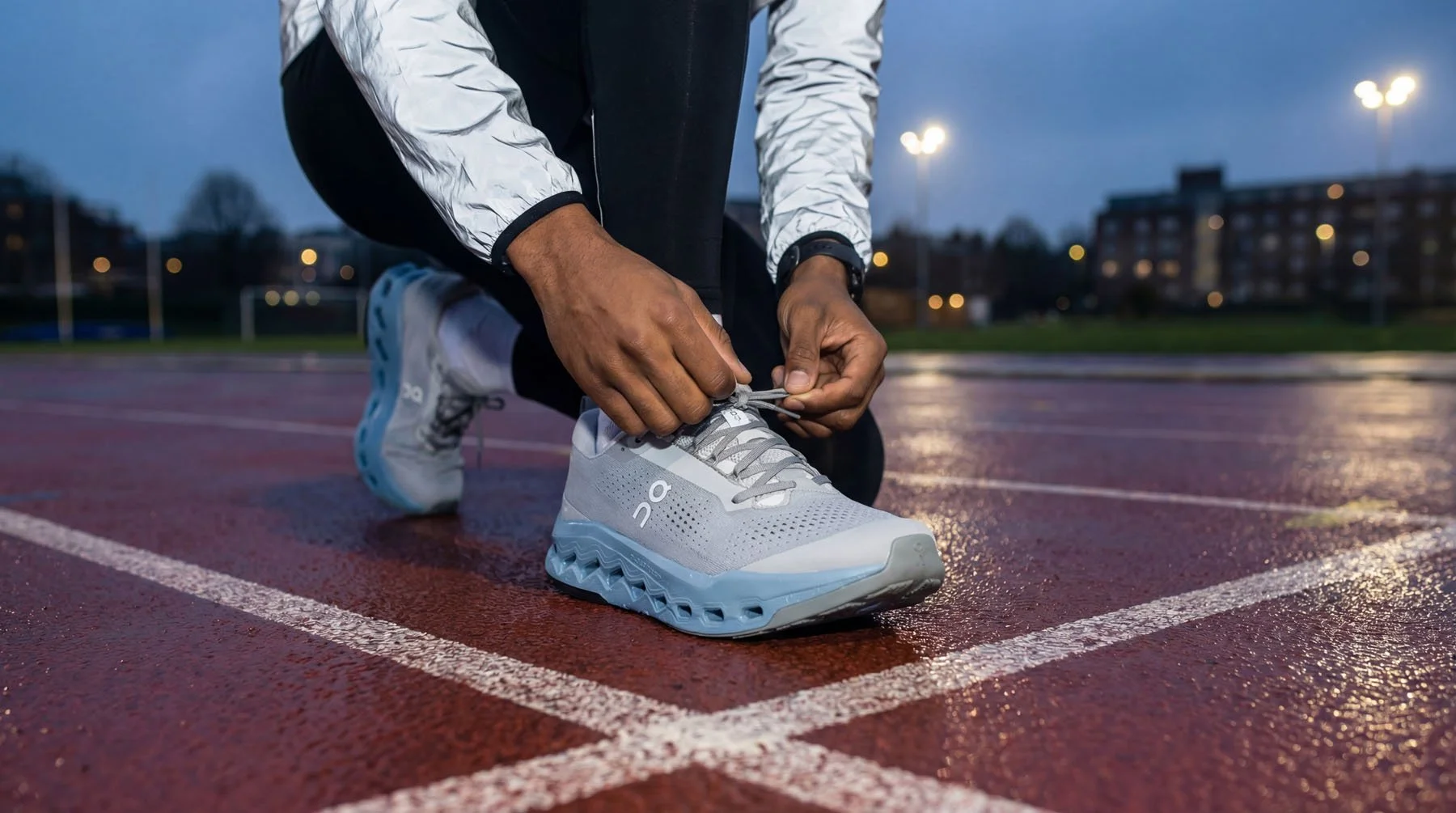 Runner tying shoelaces on a white athletic shoe on a wet outdoor track during dusk