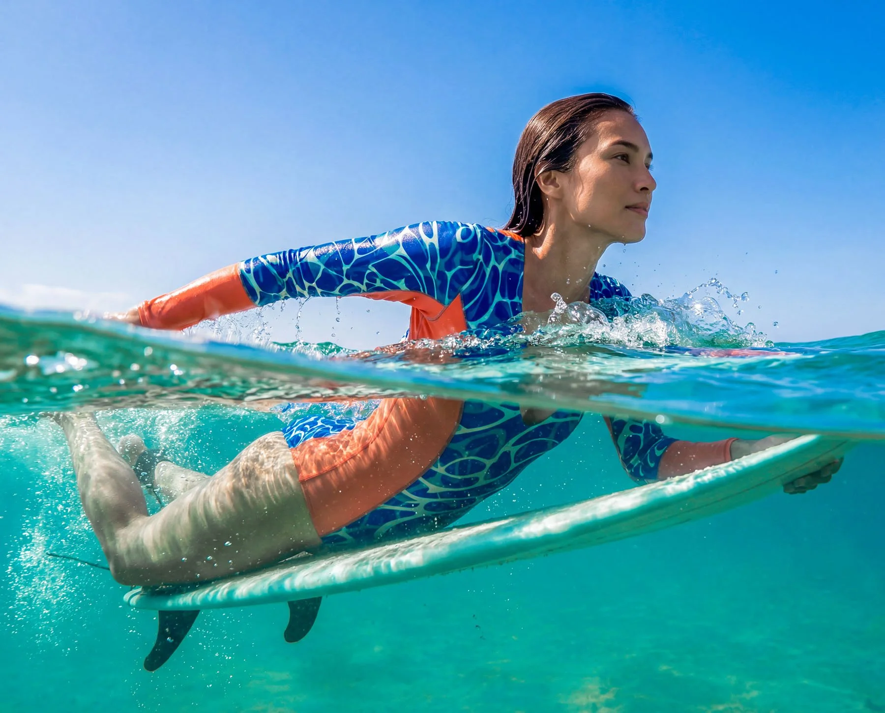 A woman in a blue and orange swimsuit paddling a surfboard in the ocean, half underwater, with a clear blue sky above.