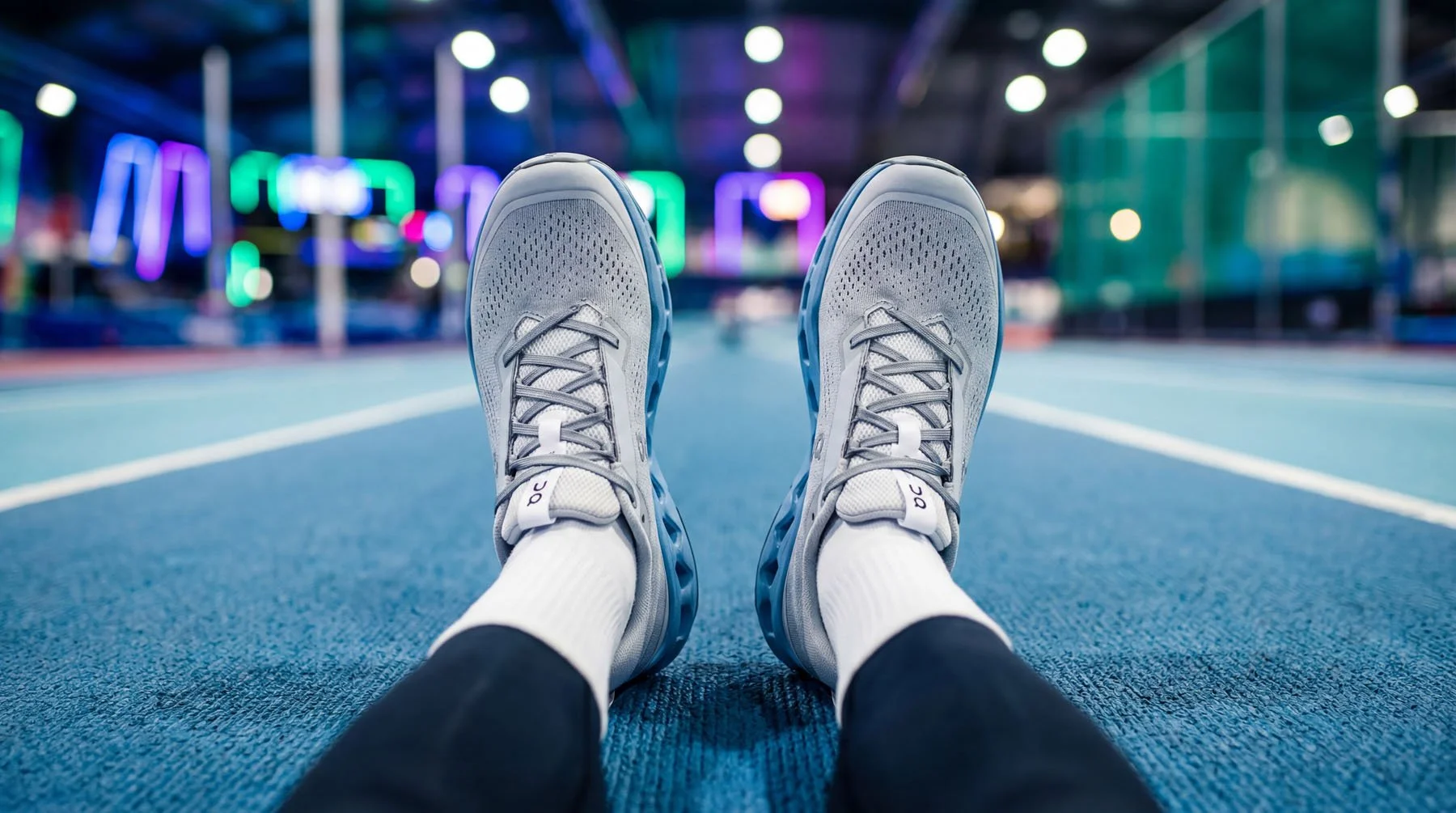 Person lying on blue indoor gym floor with legs outstretched, wearing gray athletic shoes and white socks, in front of colorful blurred gym equipment.