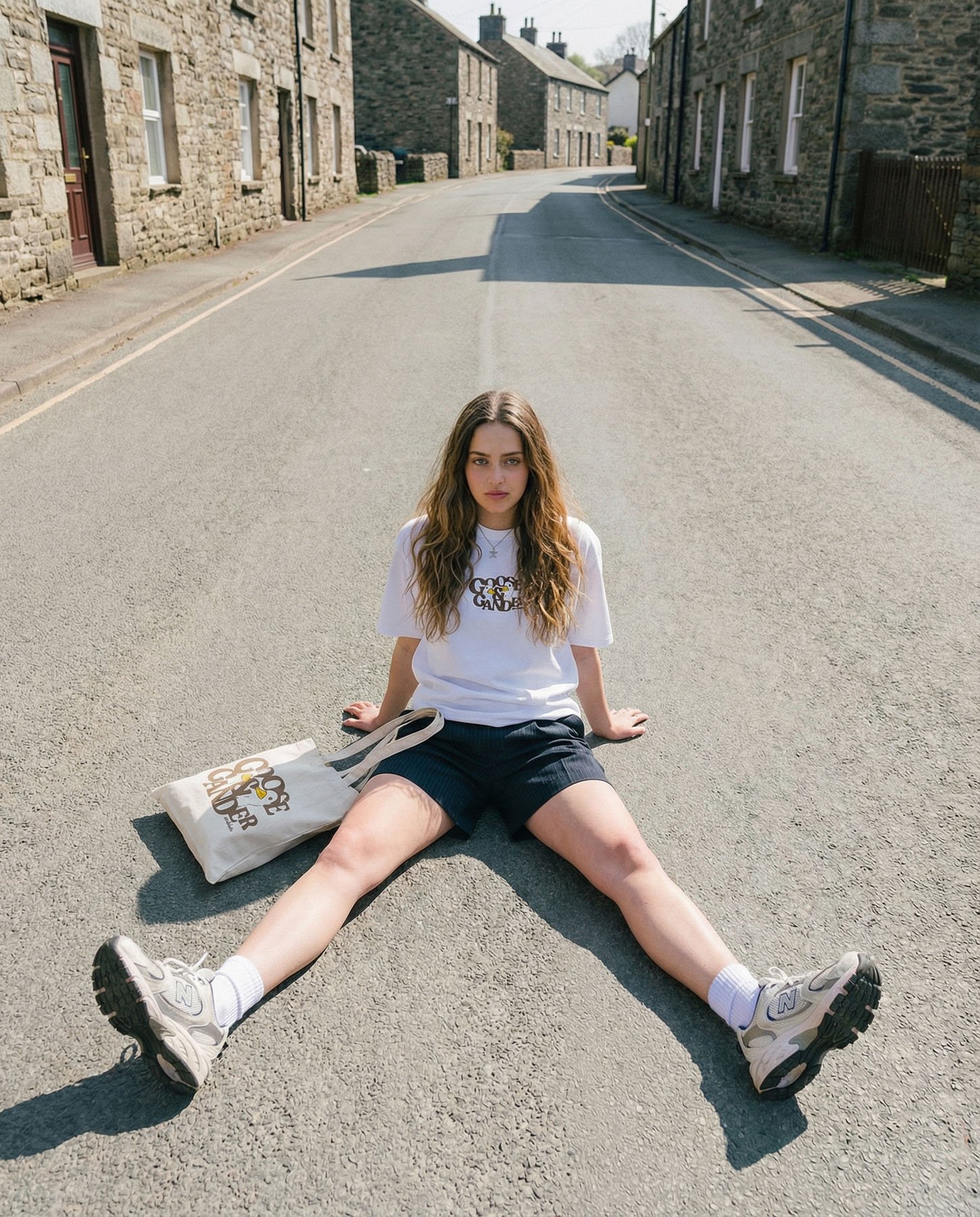 Young woman sitting on an empty street with legs spread, wearing a white t-shirt, black shorts, sneakers, and carrying a tote bag, with stone houses in the background.