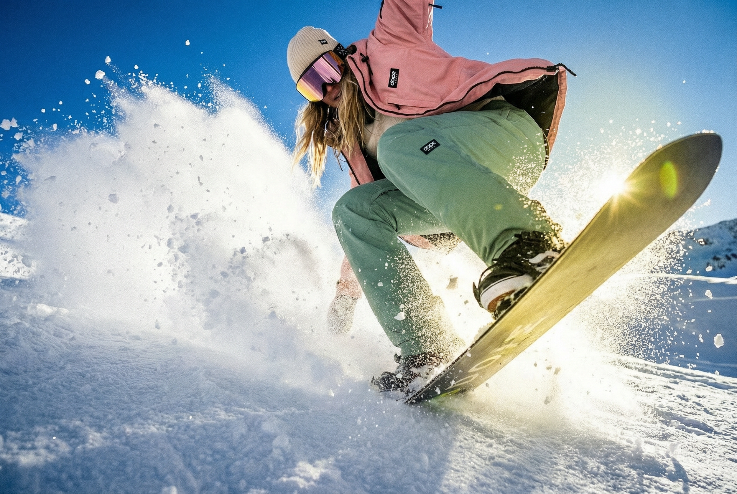 A person snowboarding in the snow wearing a pink jacket, green pants, a beige beanie, and goggles with a reflective tint, captured from a low angle with the sun shining through the snow spray.