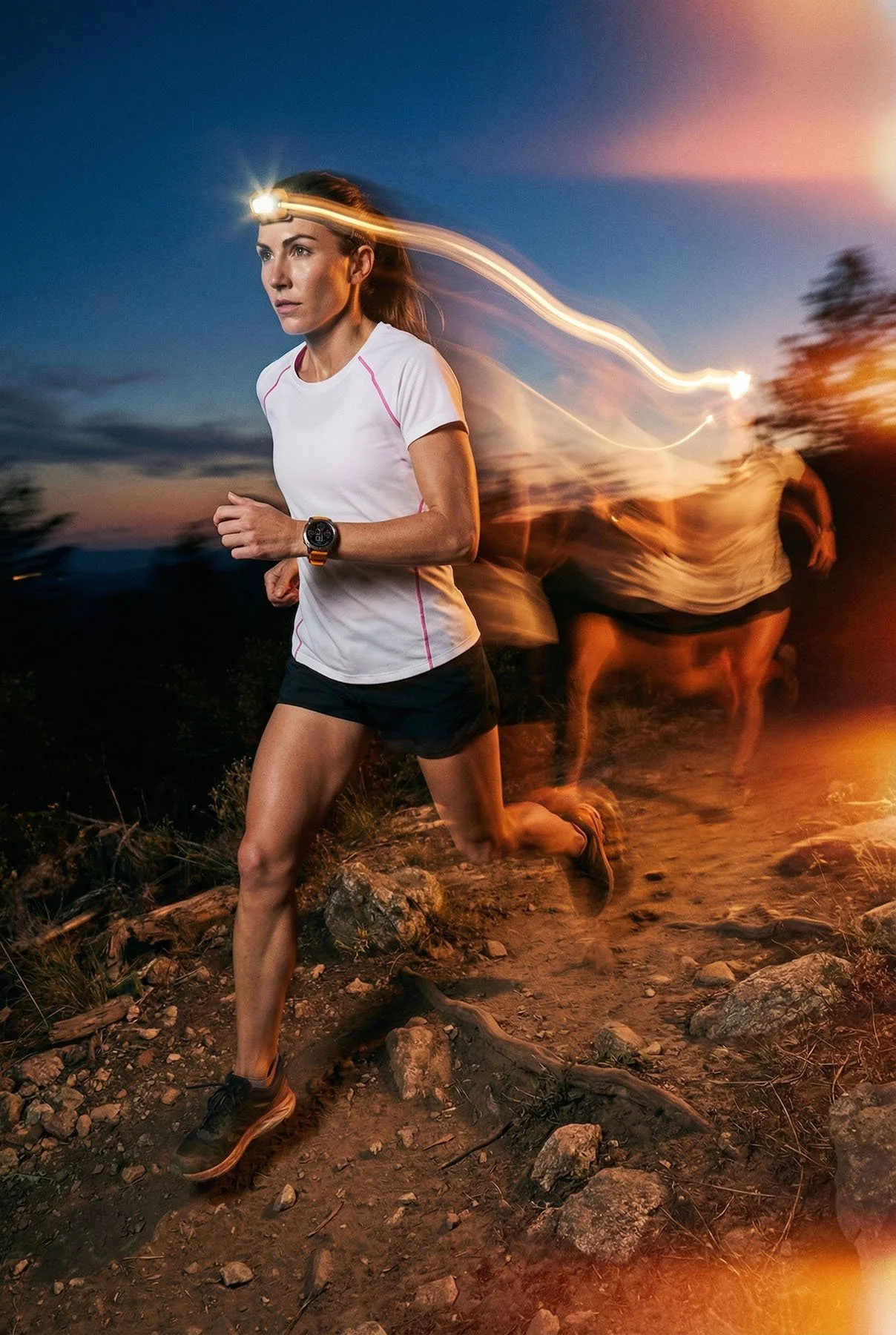 A woman running on a dirt trail at dusk or dawn. She is wearing a white athletic shirt, black shorts, a sports watch, and a headlamp. Blurred figures are running behind her.