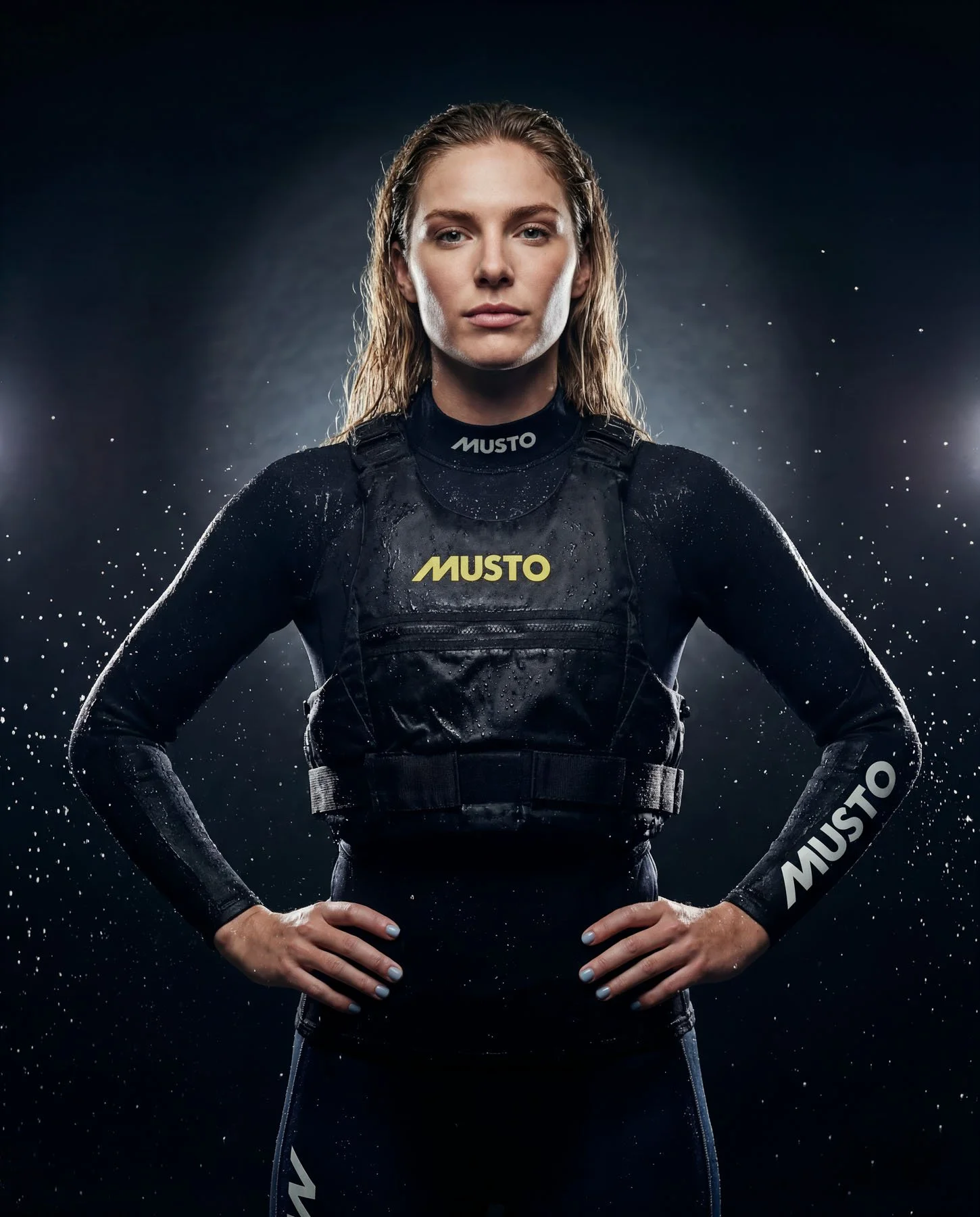 A woman in a black racing suit with wet hair, standing with hands on hips, surrounded by water droplets, and a dark background with focused lighting.
