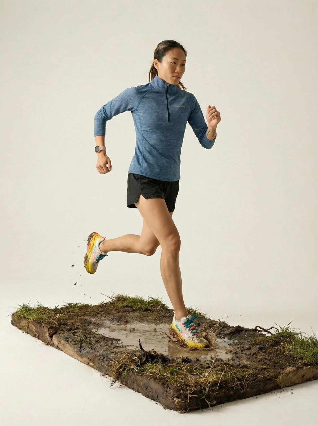 A woman running on a muddy patch of ground with water and grass, wearing a blue long-sleeve running shirt, black shorts, and colorful running shoes.