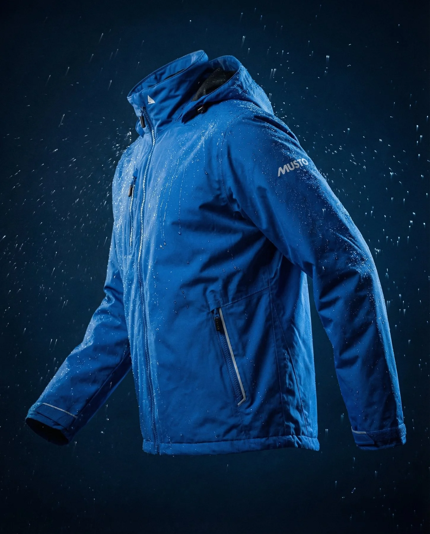 A person wearing a blue waterproof jacket with water splashing around them against a dark background.