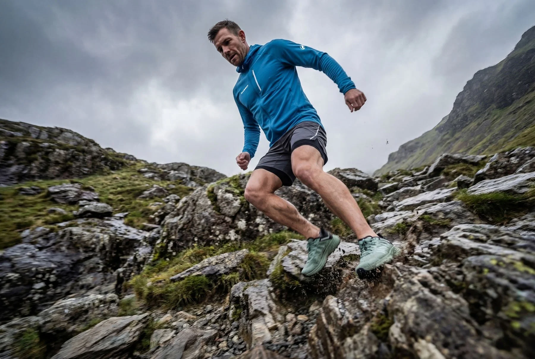 Man trail running on rocky mountain trail under cloudy sky wearing blue jacket and gray shorts