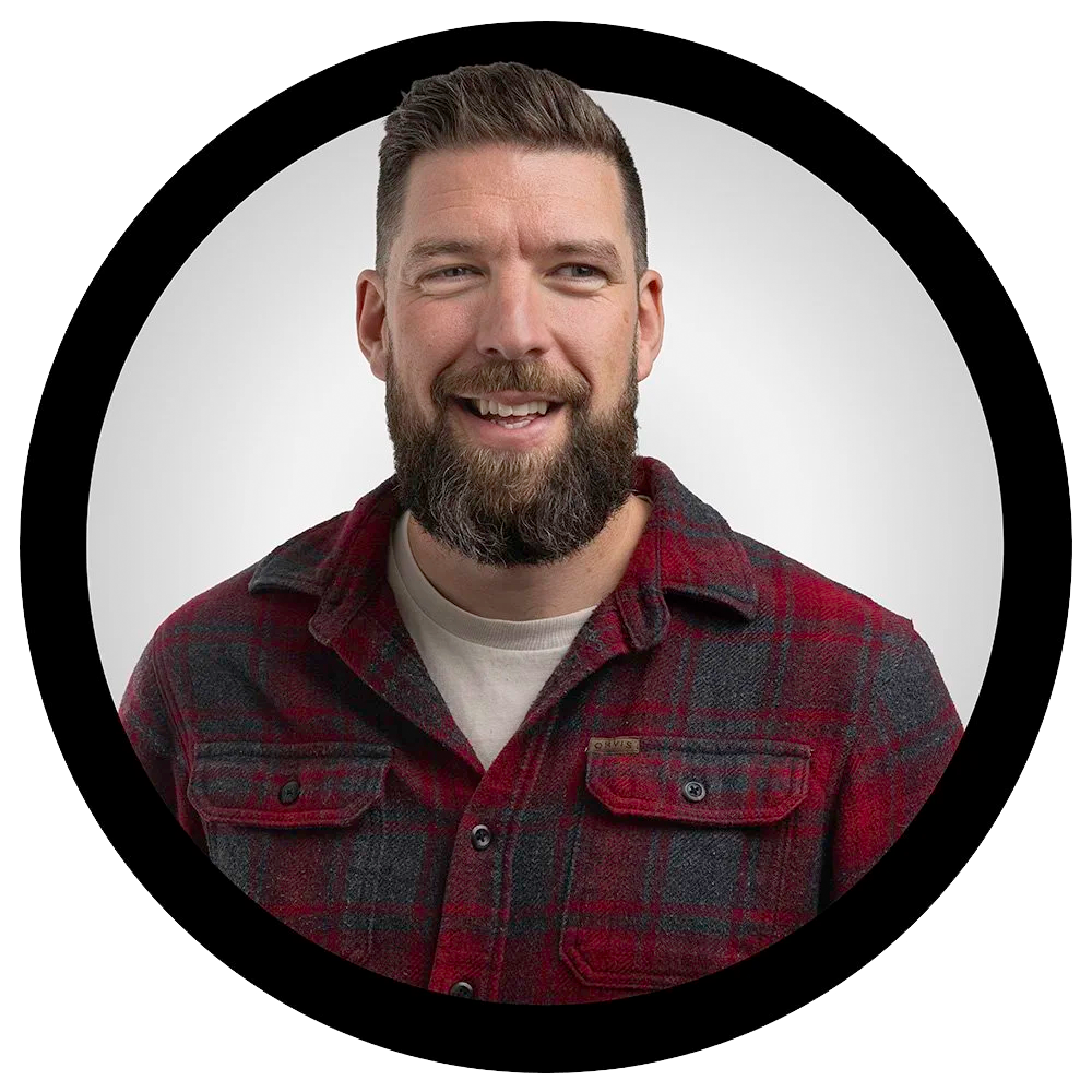A smiling man with a beard, wearing a red and gray plaid shirt, against a plain light background.