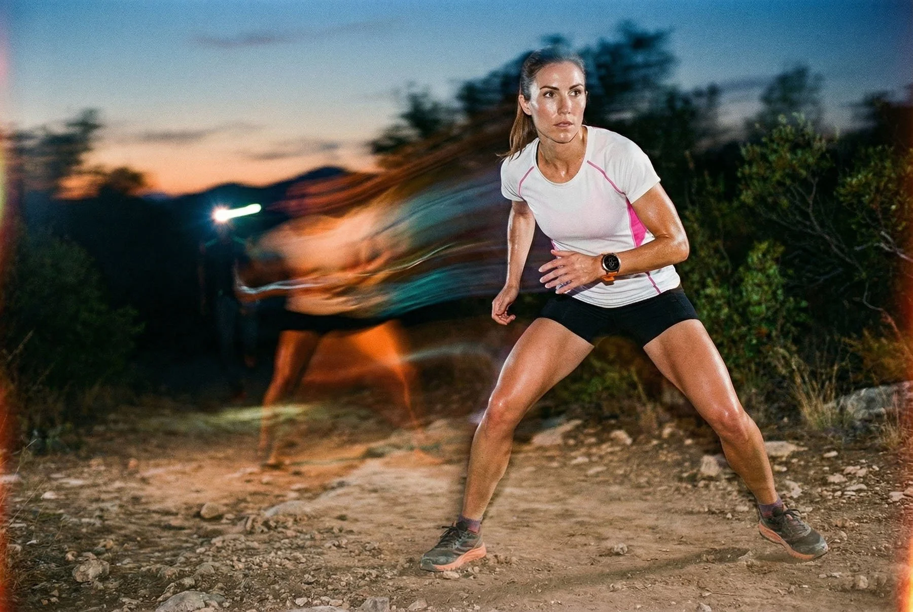 A woman in athletic clothing running on a dirt trail at sunset, with motion blur of other runners in the background.