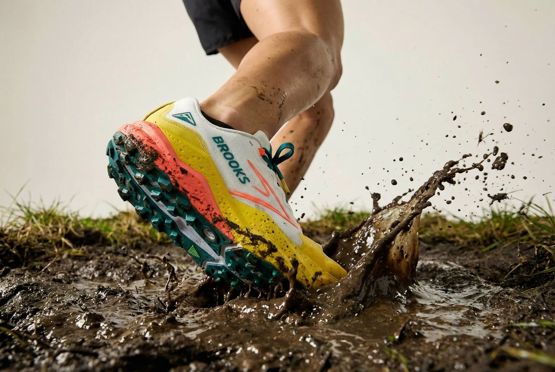 Close-up of a person running through muddy ground, wearing a yellow, white, and teal Brooks trail running shoe with mud on the sole, splashing muddy water.