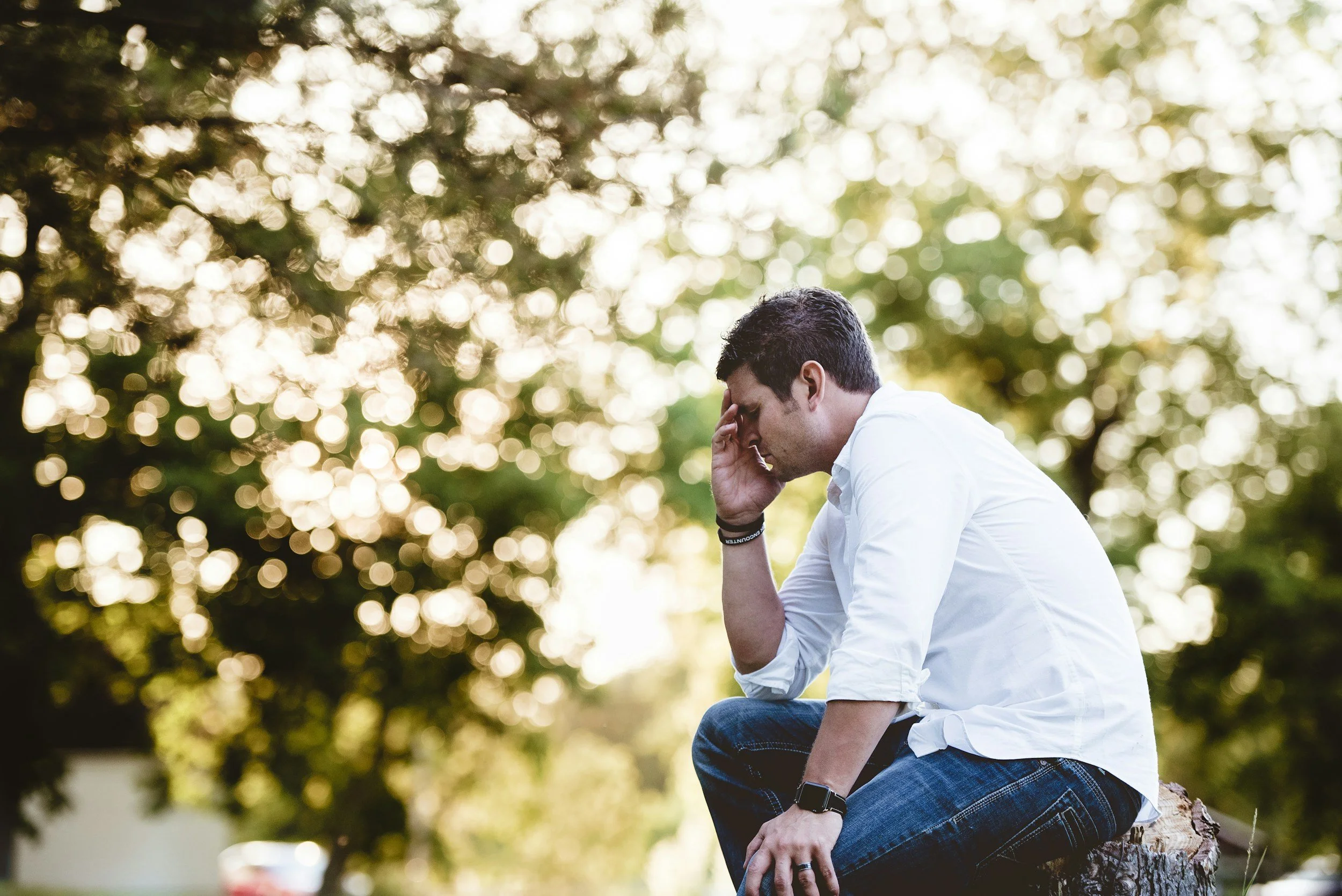 A man sitting on a log outdoors with his hand on his forehead, looking down, with blurred trees and sunlight in the background.