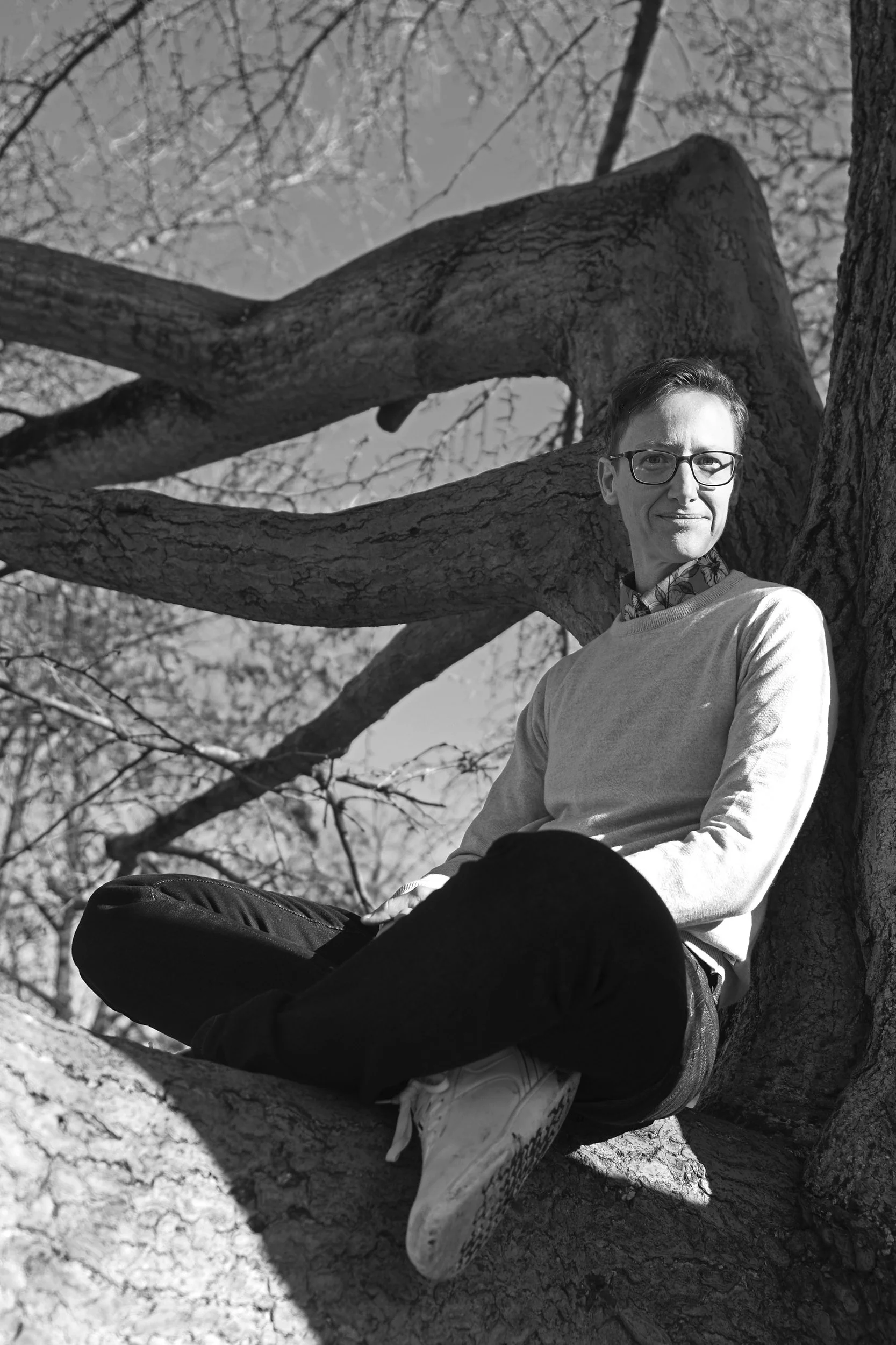 Schwarz-weiß-Foto eines Mannes, der auf einem Baum sitzt, mit Brille, heller Kleidung und dunklen Hosen, im Freien bei Tageslicht, hinter ihm Baumzweige.