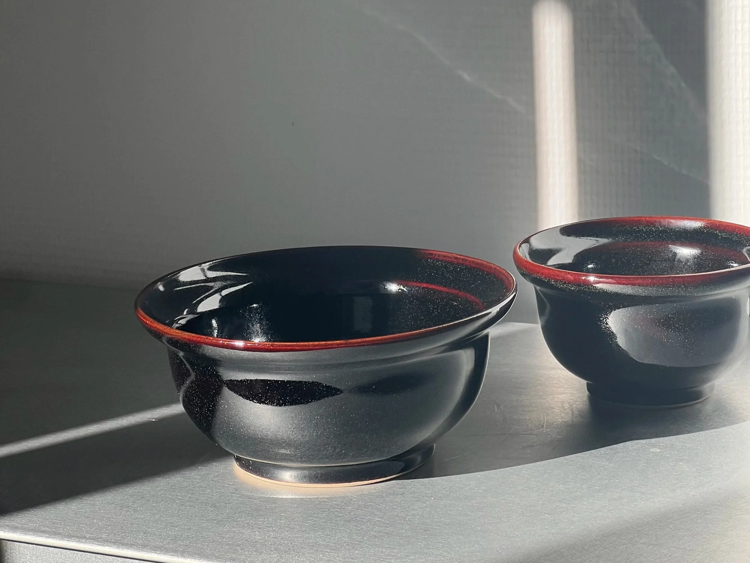 Deux bols noirs avec intérieur rouge, en céramique, placés sur une surface plane métallique.
