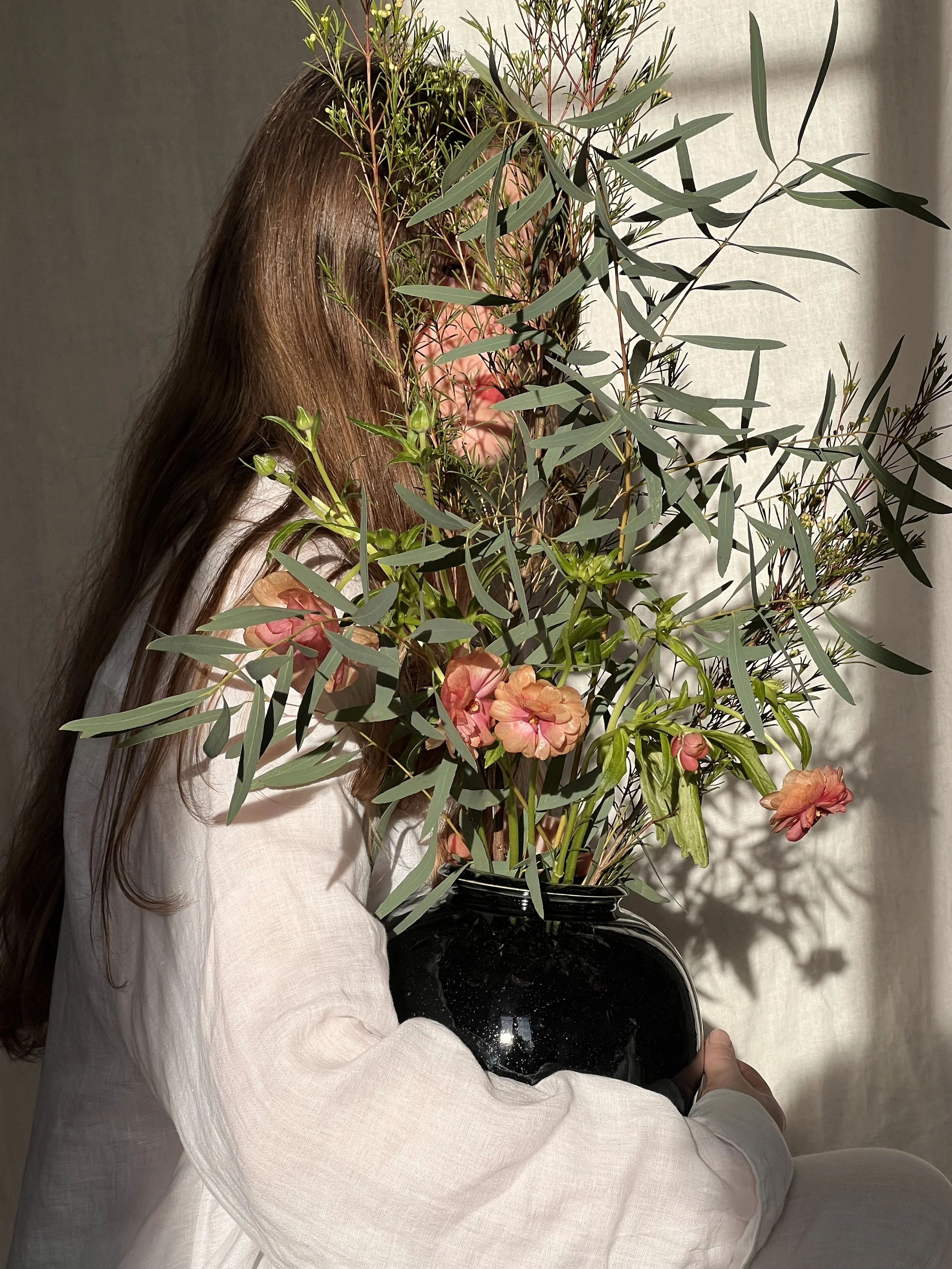 Une personne habillée en blanc tient un vase noir avec un arrangement floral composé de feuillage vert et de fleurs roses. La personne est vue partiellement de côté, et le fond est clair. La composition donne une impression artistique et élégante.