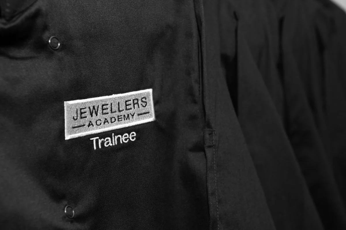 Close-up of a black jacket with a patch that reads "Jewellers Academy" and a smaller text "Trainee" underneath.