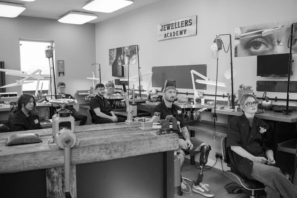 People sitting in a jewelry-making workshop at Jewelers Academy, with workbenches, tools, and jewelry displays on the walls.