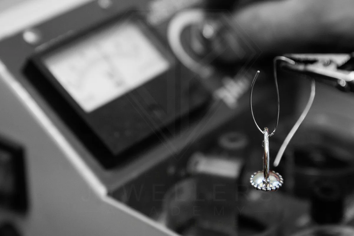 A silver hoop earring hanging from a hook with a small pearl in the center, blurred cash register and other objects in the background.