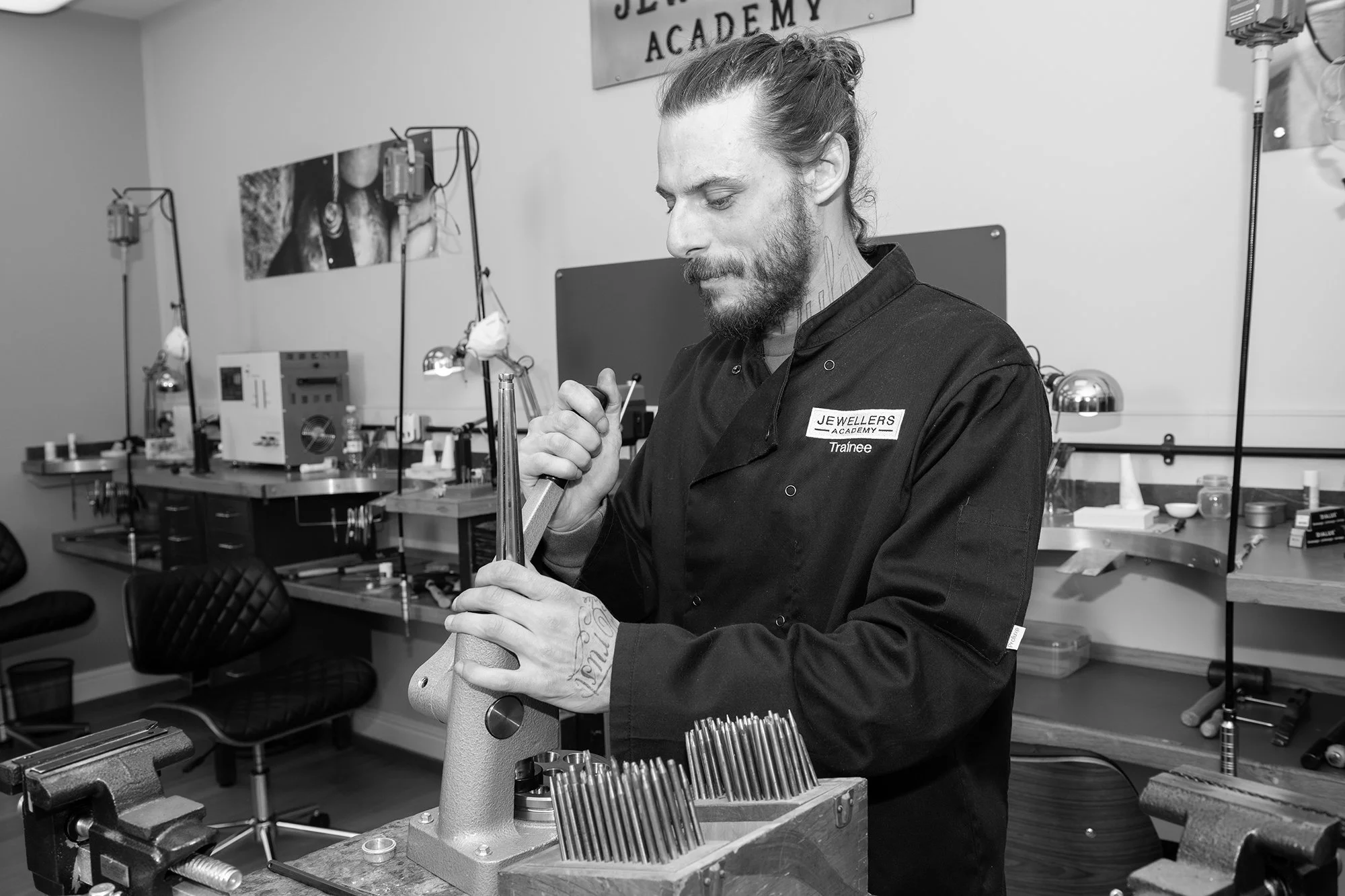 Black and white photo of man with long hair and beard, wearing a jacket labeled 'Jewellers Academy Trainee,' working on a jewelry piece at a workbench in a jewelry workshop.