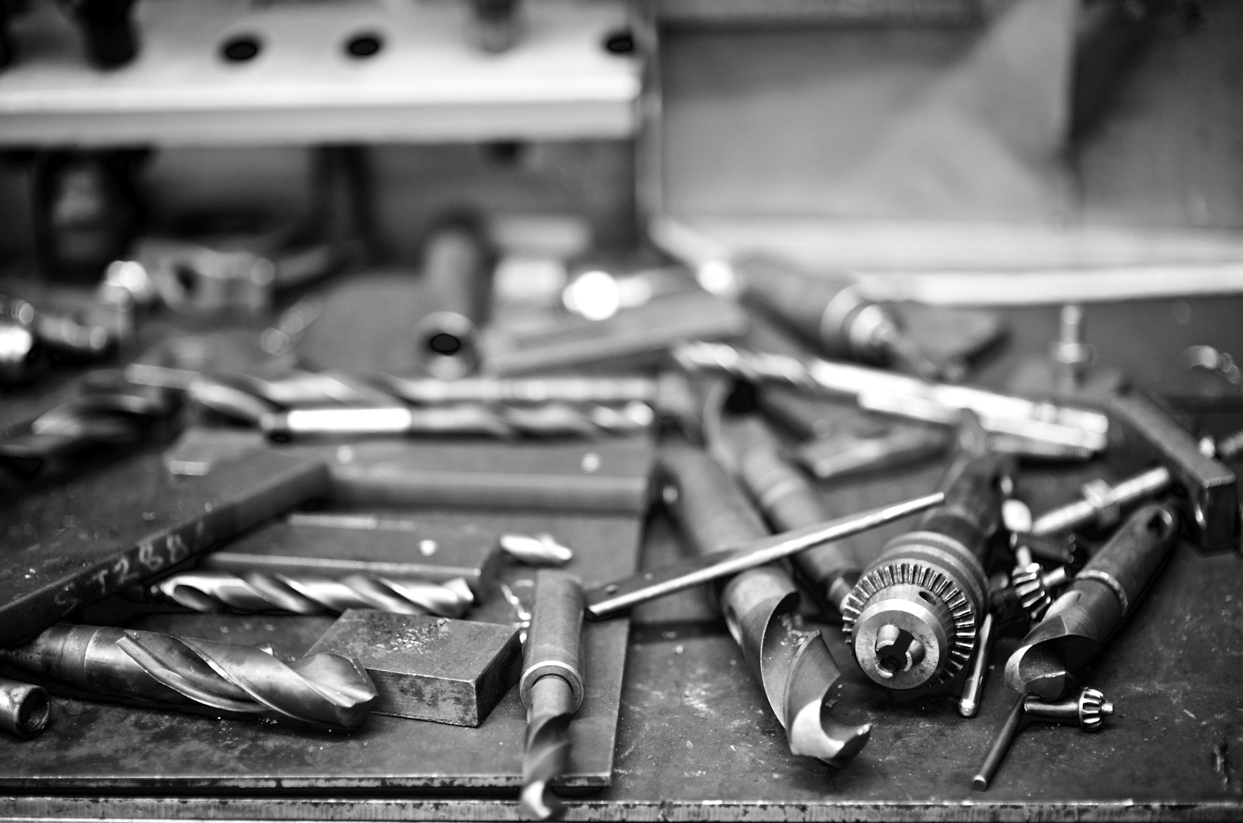 A cluttered workbench with various metal tools and parts, including drill bits, gears, and screws, in black and white.