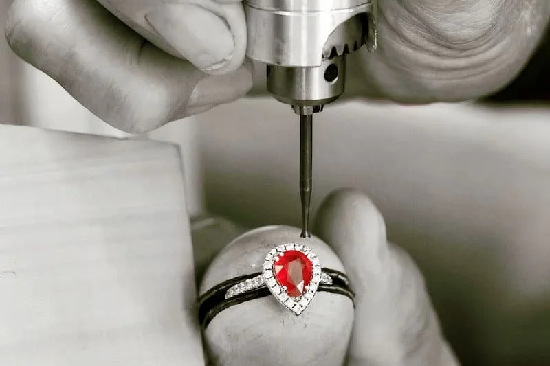 Close-up of a jewelry artisan setting a large red gemstone ring with a diamond halo using a precision tool.