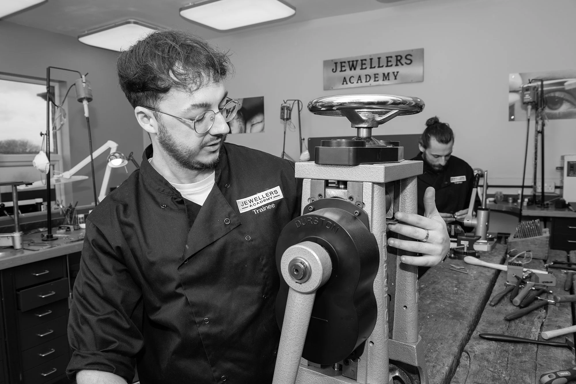 A man with glasses and a beard operating a jewelry making machine in a workspace labeled 'Jewellers Academy'. He is focused on the machine while another person works in the background.